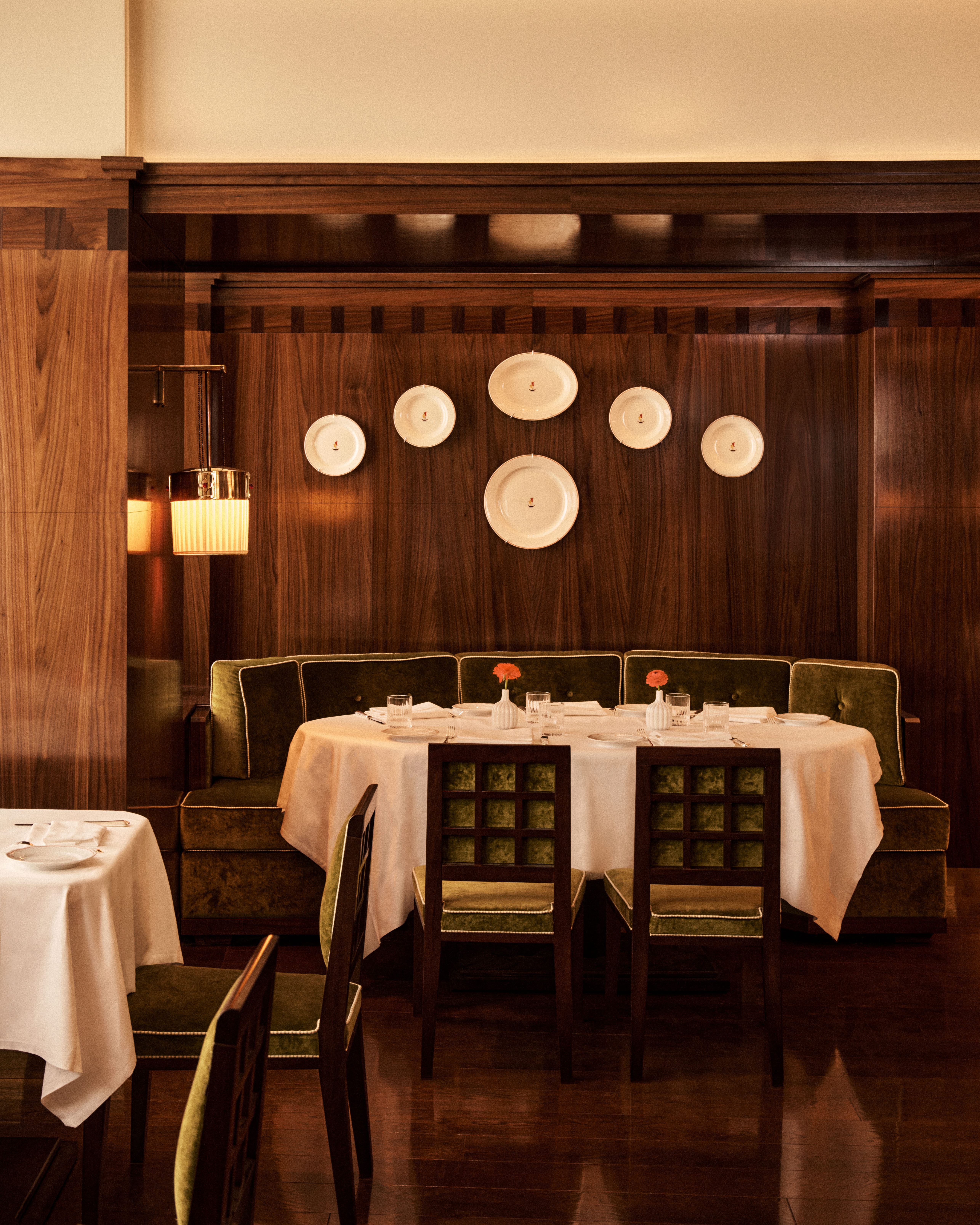 Elegant banquette dining area with rich walnut paneling, green velvet seating, white tablecloth–draped tables, sculptural wall-mounted plates, and warm ambient lighting creating a classic, intimate restaurant atmosphere.