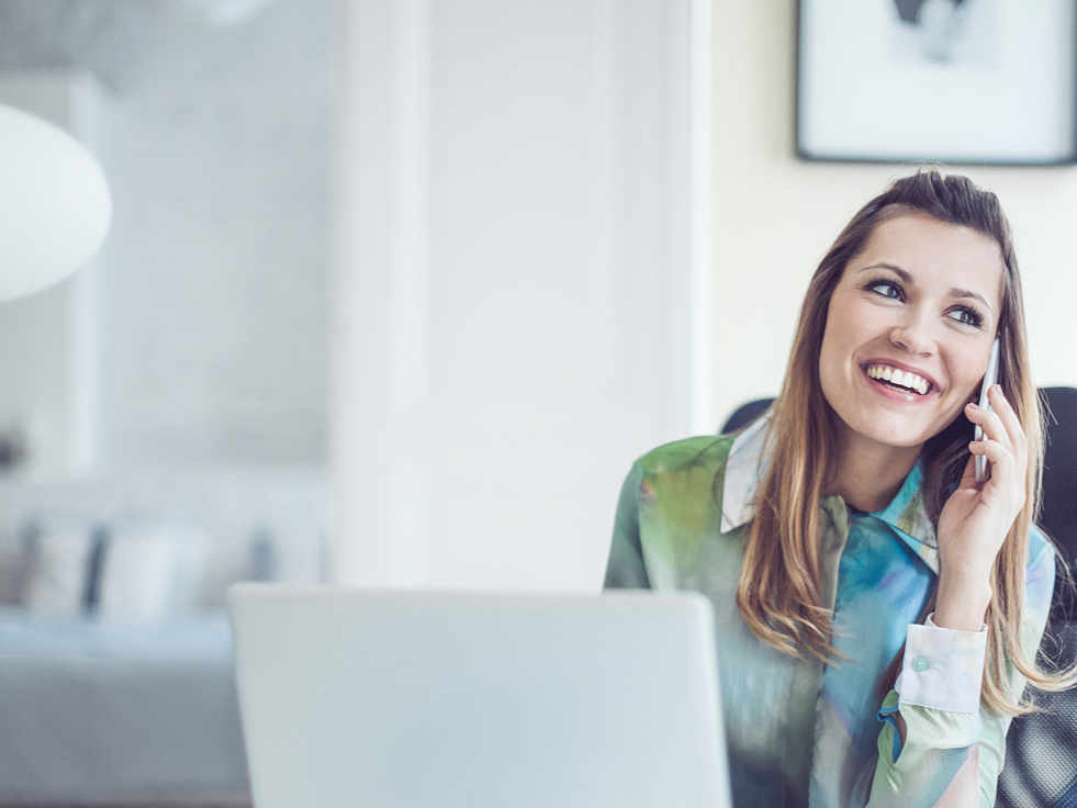 A woman in a colorful blouse smiles while talking on her phone at a desk. A blurred background with a framed picture adds to the casual atmosphere.
