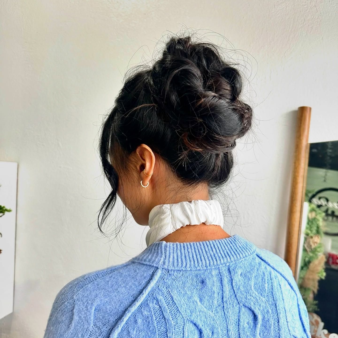 Woman with dark hair styled in messy bun updo with loose strands, photographed from back in soft indoor setting.