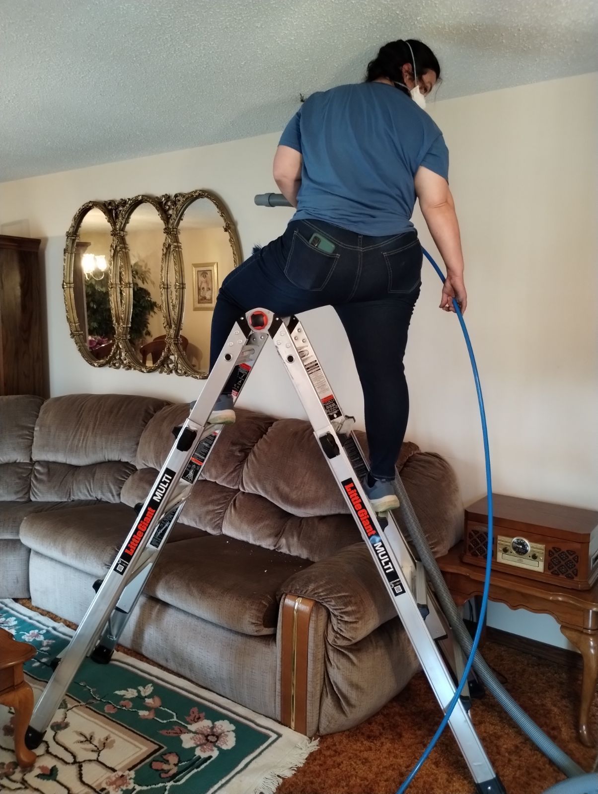 Technician standing on ladder inside living room, cleaning air ducts near ceiling using hose equipment, with couch and decor visible, demonstrating residential HVAC maintenance and indoor air quality service.