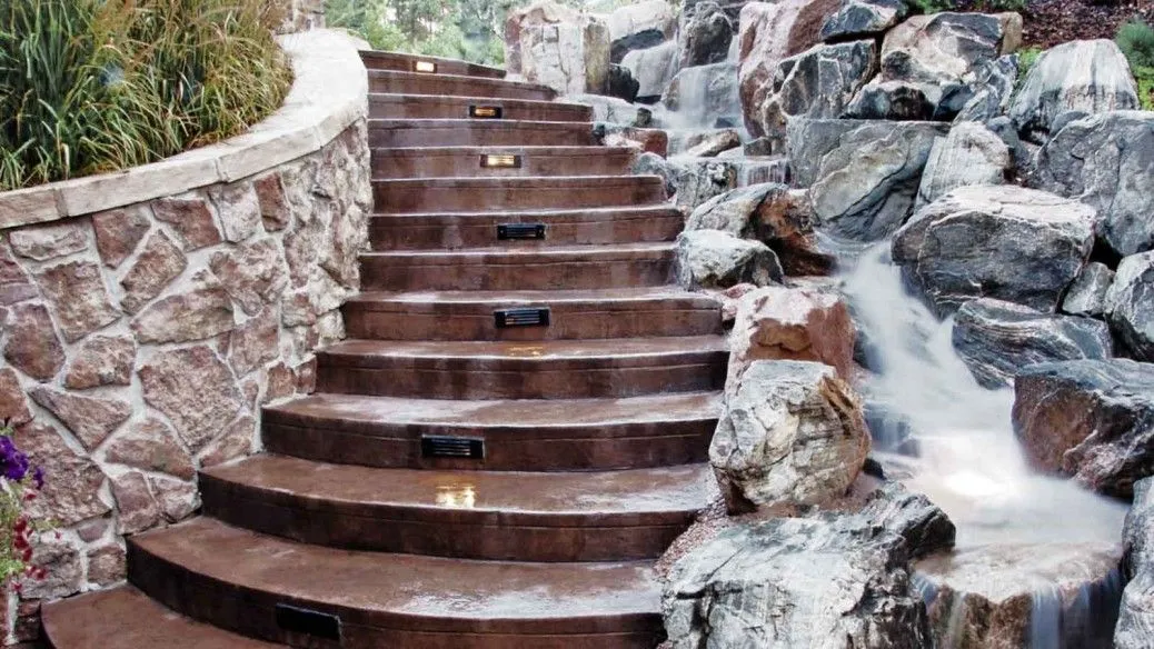 A curved, brown concrete staircase with integrated lighting ascends alongside a rock formation with a cascading waterfall. The staircase is bordered on the left by a stacked stone retaining wall topped with lush greenery. The waterfall flows over large, grey and brown rocks into a pool at the base. The scene appears to be an outdoor landscape feature.