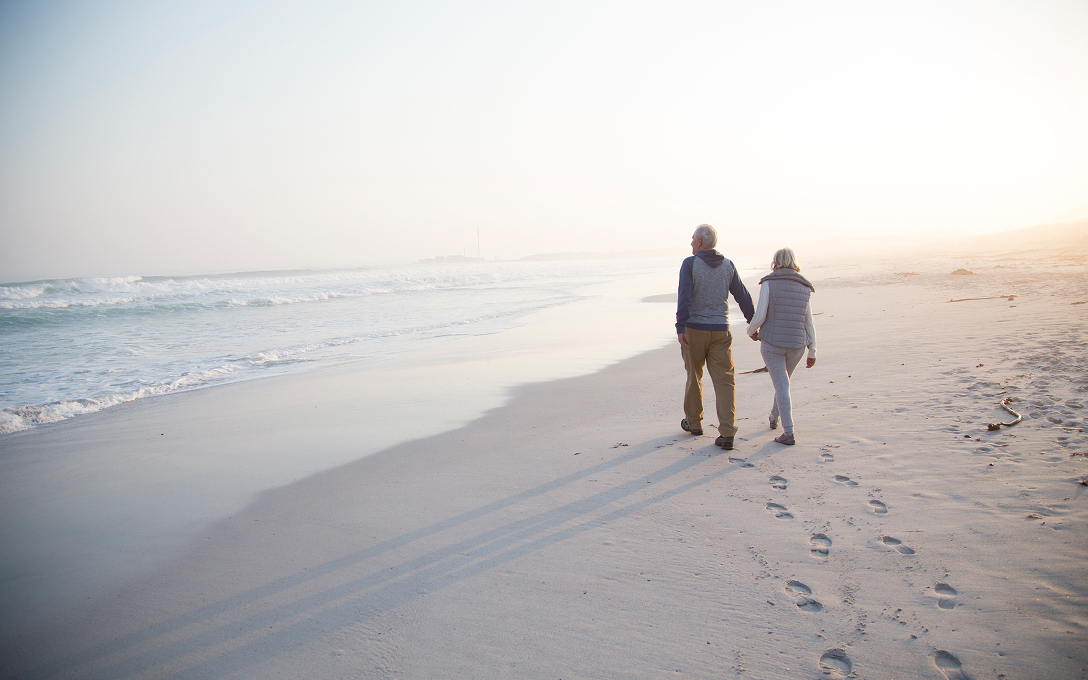 An elderly couple walks hand in hand along a serene beach at sunrise, casting elongated shadows on the sand. Waves gently lap the shore, creating a peaceful atmosphere.