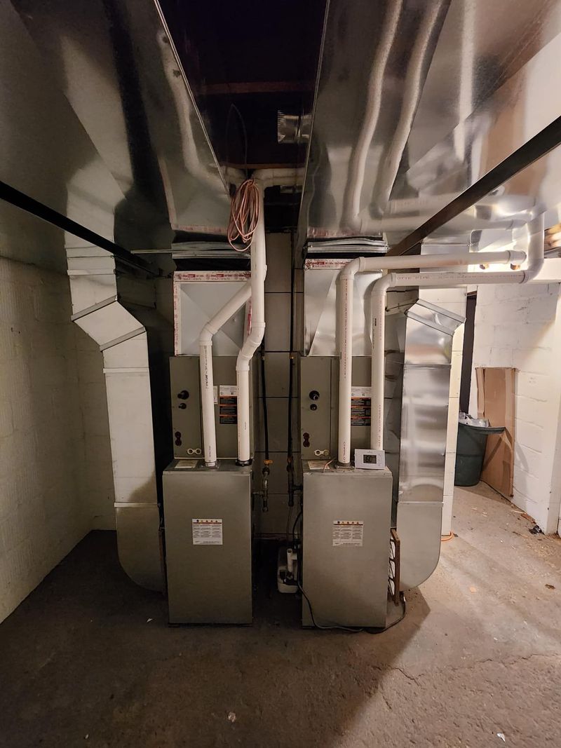 Two identical silver furnaces are installed side-by-side in a basement, connected to an extensive network of shiny metal ductwork and white PVC venting pipes for efficient home climate control.