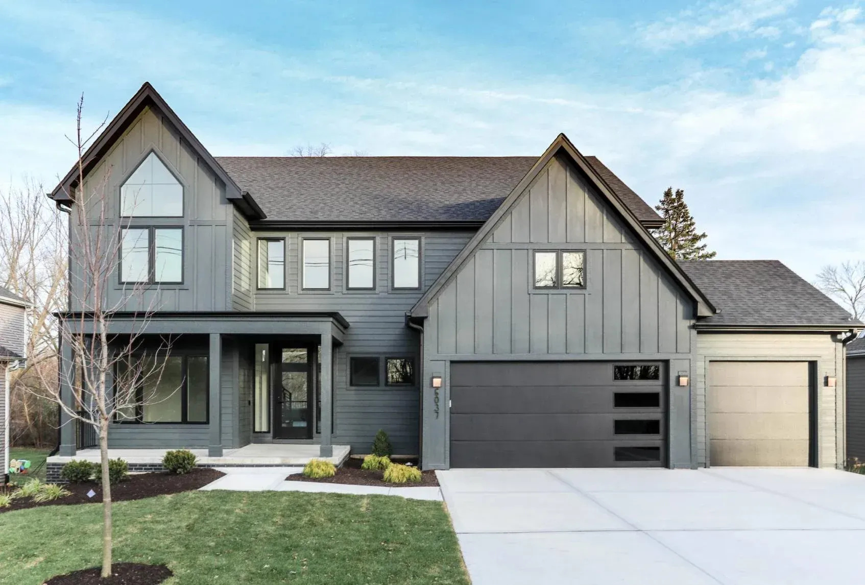 A contemporary gray home features a charcoal garage door with horizontal windows beside a cream-colored single garage door, complemented by modern landscaping and a broad driveway with clean lines today.