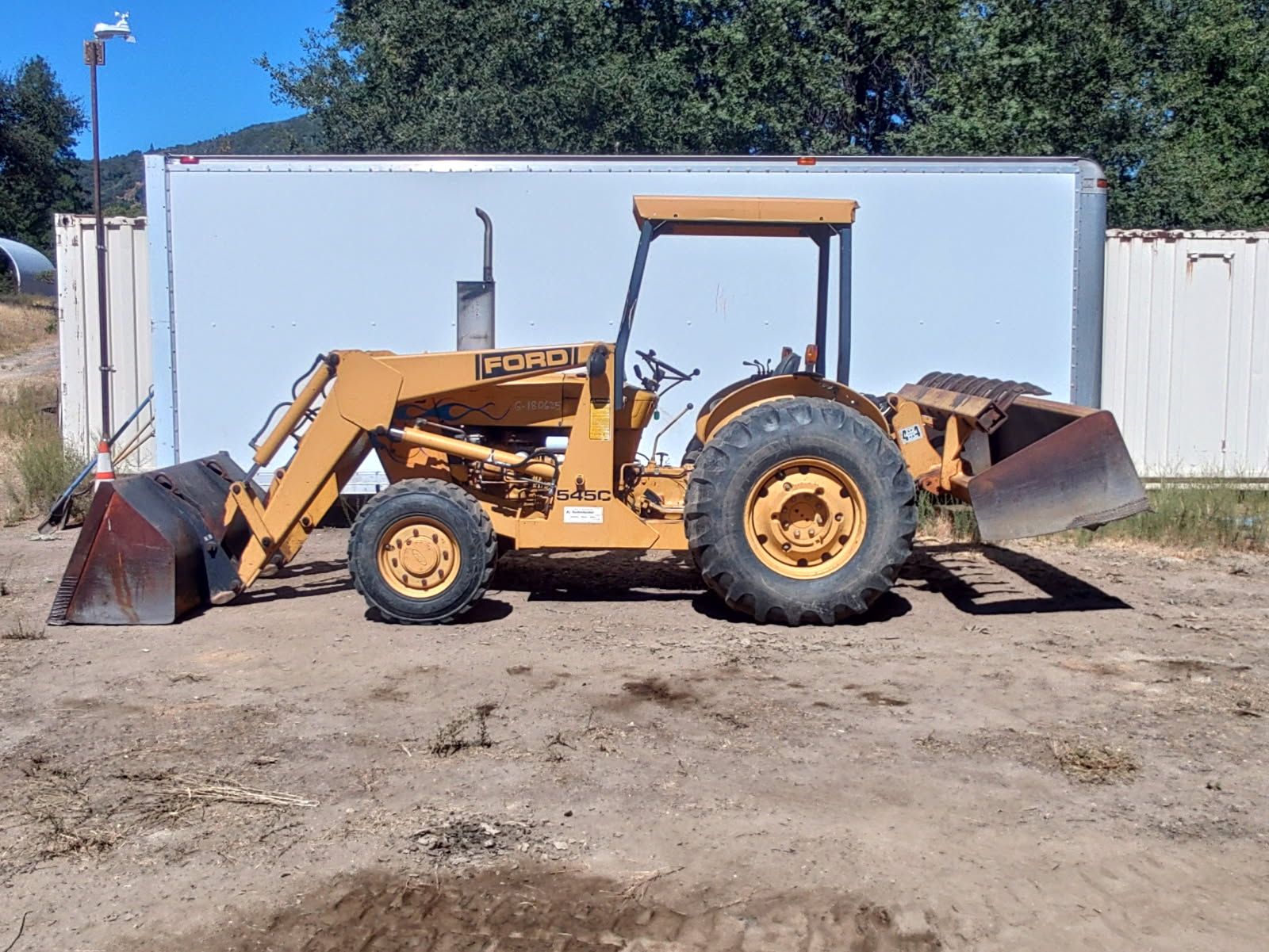 Yellow Ford tractor-loader is parked on dirt beside a large white enclosed trailer, featuring a front bucket, rear box blade, large rear tires, open operator station, and hydraulic loader arms.