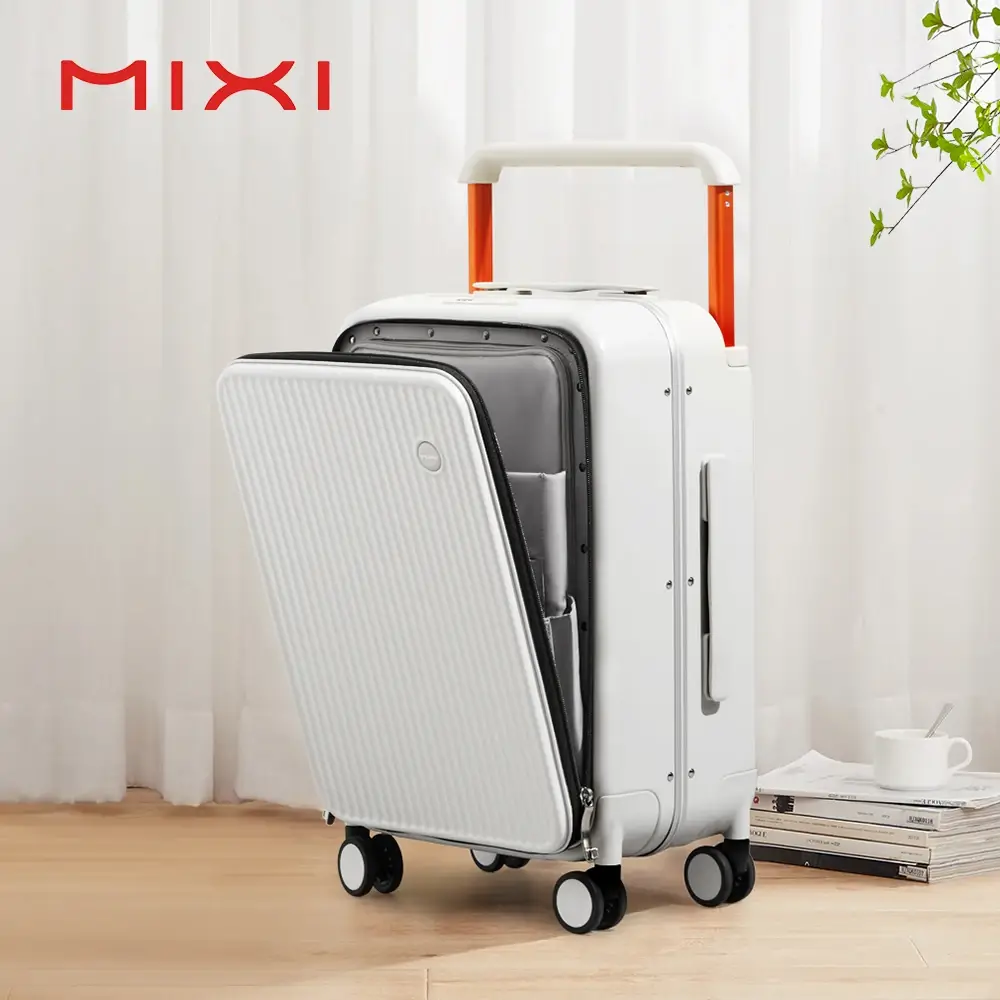 A white MIXI suitcase with orange accents and a partially opened front compartment sits on a wooden floor. The suitcase has a retractable handle and four wheels. A stack of books and a white mug are placed to the right of the suitcase. The background is a light-colored curtain.