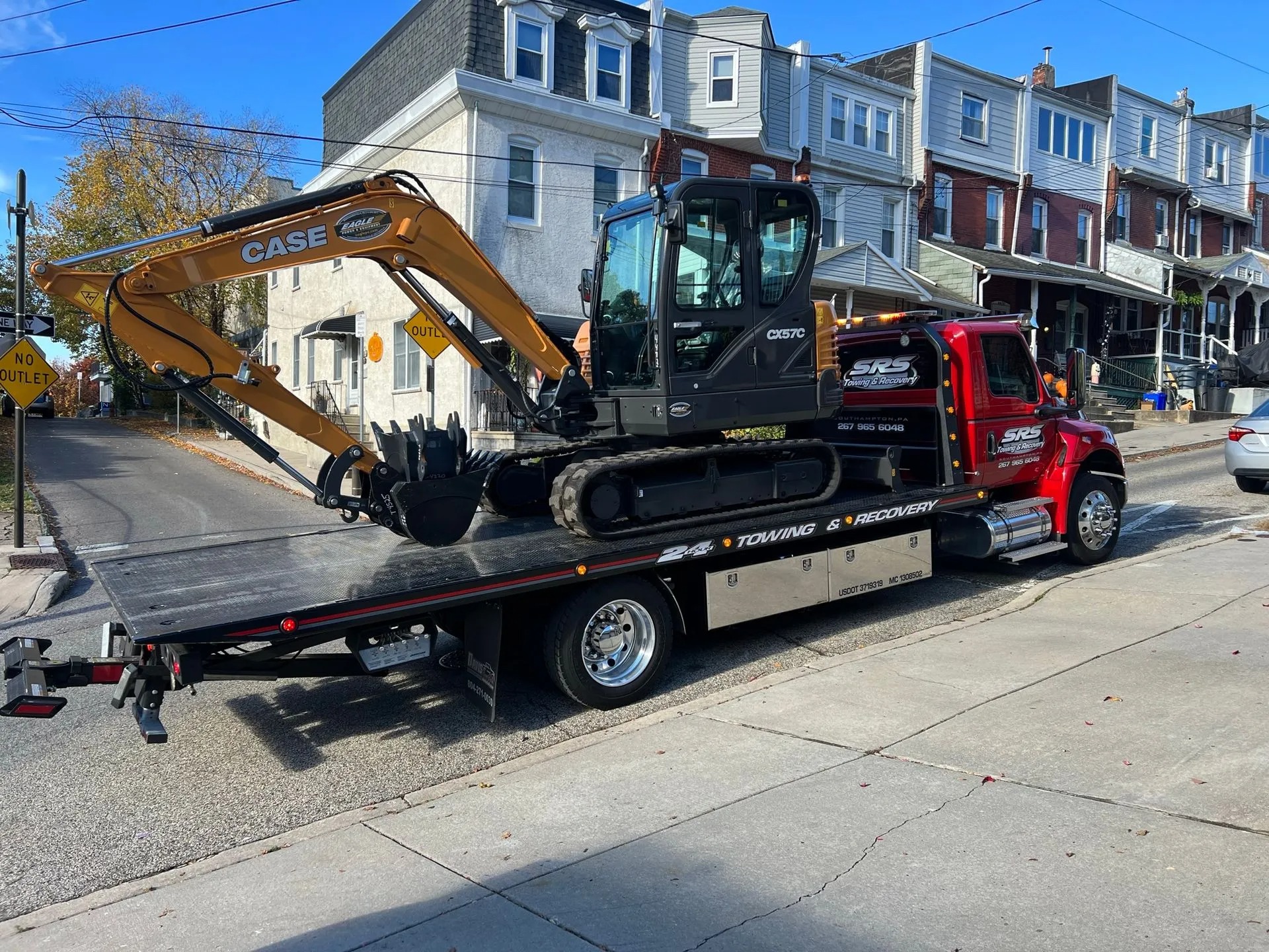 SRS Towing & Recovery’s red flatbed tow truck carrying a CASE CX57C excavator, parked on a street lined with row houses.