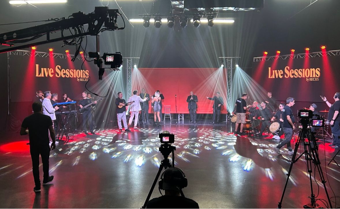Aqu&iacute; capturamos una vista panor&aacute;mica de nuestro escenario principal durante la grabaci&oacute;n en vivo de una orquesta completa. Se observa a los m&uacute;sicos distribuidos en la tarima y un imponente despliegue de luces blancas y rojas que reflejan patrones geom&eacute;tricos en el suelo. Al frente y en los laterales, se aprecian nuestras c&aacute;maras profesionales montadas en tr&iacute;podes y gr&uacute;as de filmaci&oacute;n. Nos encanta transformar nuestro ballroom en el mejor sal&oacute;n de eventos de la ciudad, ideal si buscas salones para show de alto nivel.