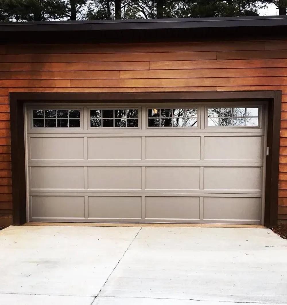 A traditional white sectional garage door features rectangular recessed panels and a row of upper windows, framed by dark wood siding and a clean concrete driveway for balanced curb appeal.