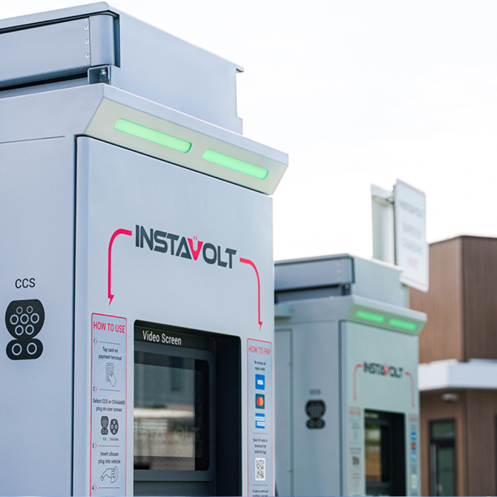 Close-up of an InstaVolt electric vehicle charging station with glowing green status lights, payment instructions, and charging connectors visible. Another charger is in the background outside a modern building.