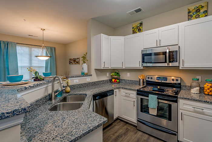 A kitchen with granite countertops, stainless steel appliances, hardwood-style flooring, a double basin sink, and white cabinets near a dining room.