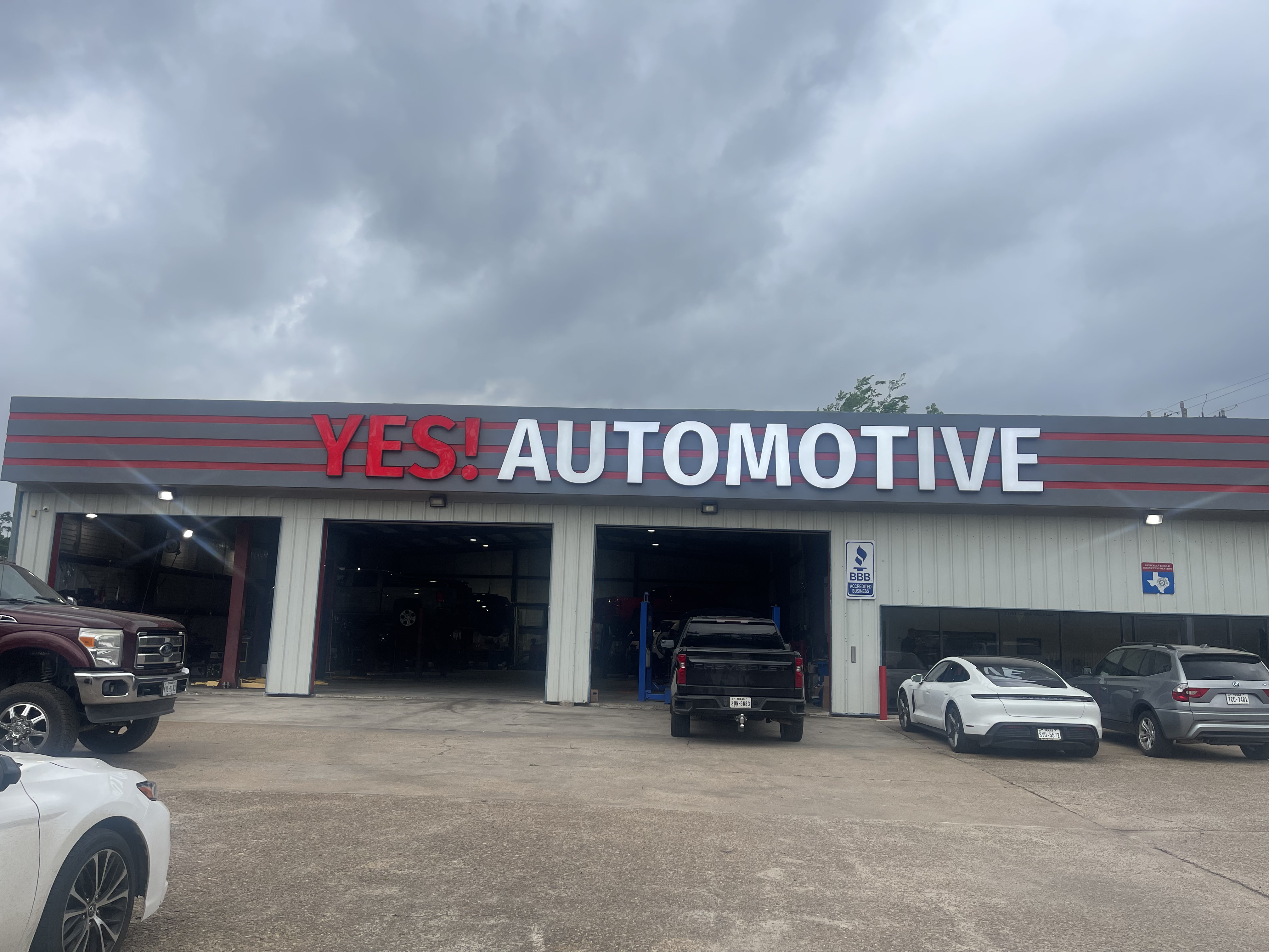 Front exterior view of YES! Automotive, Porter, TX, a full-service auto repair and transmission repair shop. The building features prominent red and white YES! AUTOMOTIVE signage, multiple open service bays, and customer vehicles parked in front. The facility displays a BBB Accredited Business sign and provides a spacious parking area with convenient access to automotive repair and maintenance services.
