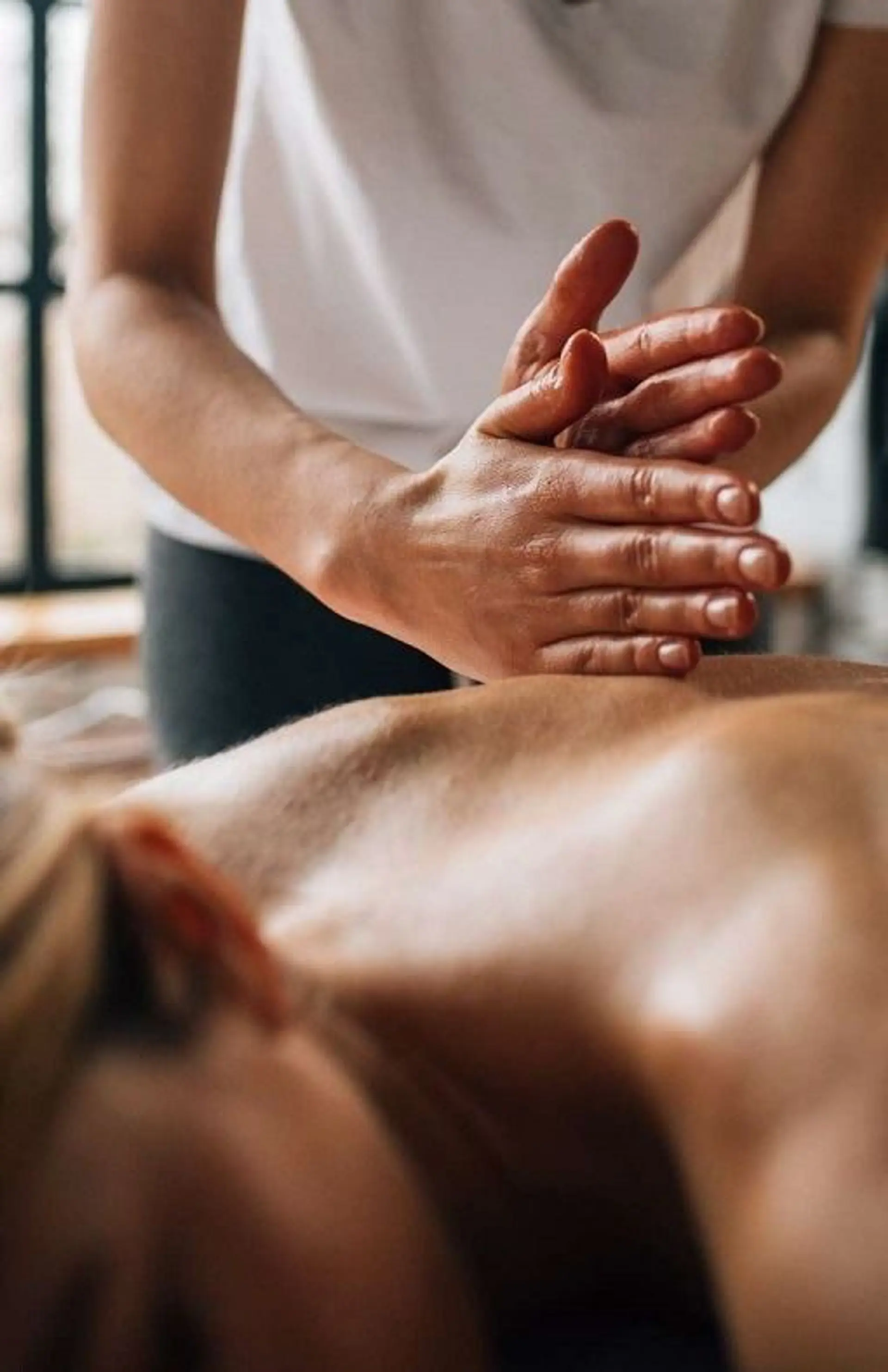 A close-up shot shows a person's hands massaging another person's back, with oil visible on the hands. The background is blurred and suggests a spa or wellness setting with natural light.