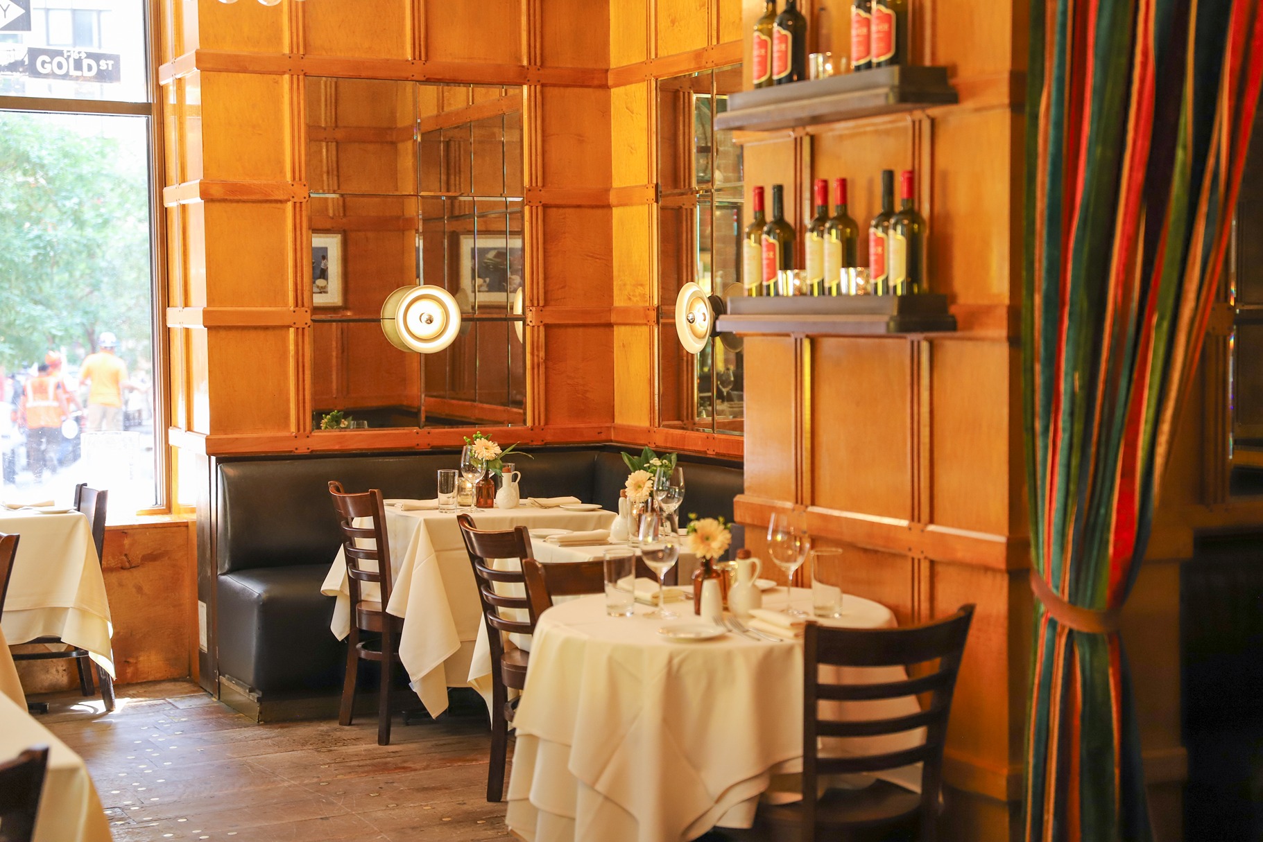 Warm sunlight streams onto white-clothed tables and leather banquettes. The space features light wood paneling, mirrored accents, and a vibrant striped curtain tied to the side.