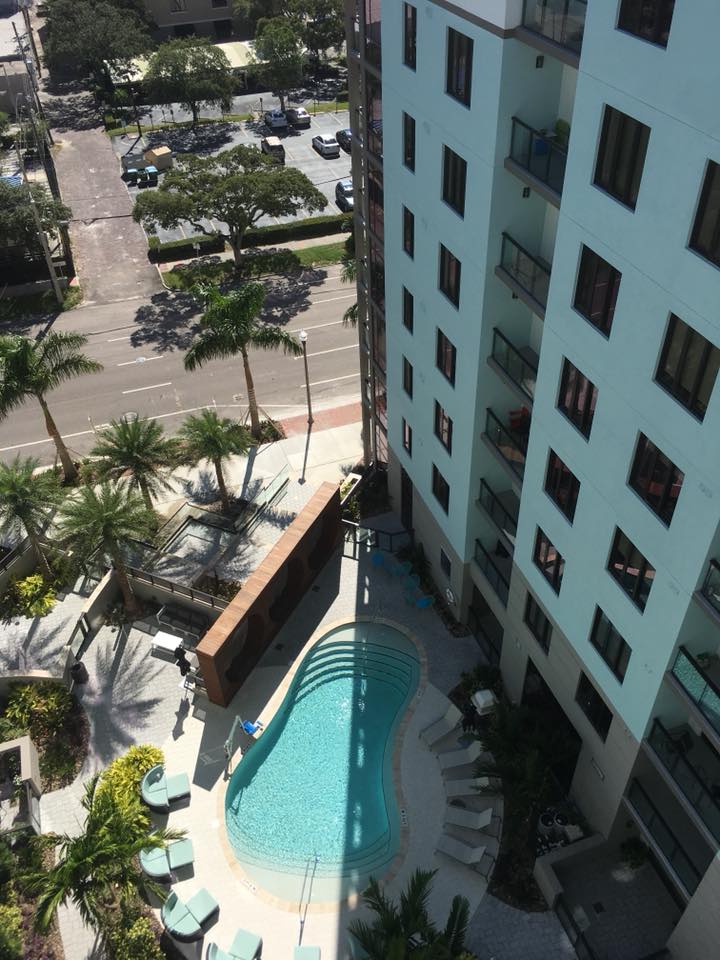 Aerial view of a commercial or condominium swimming pool surrounded by a high-rise building, palm trees, and poolside seating.