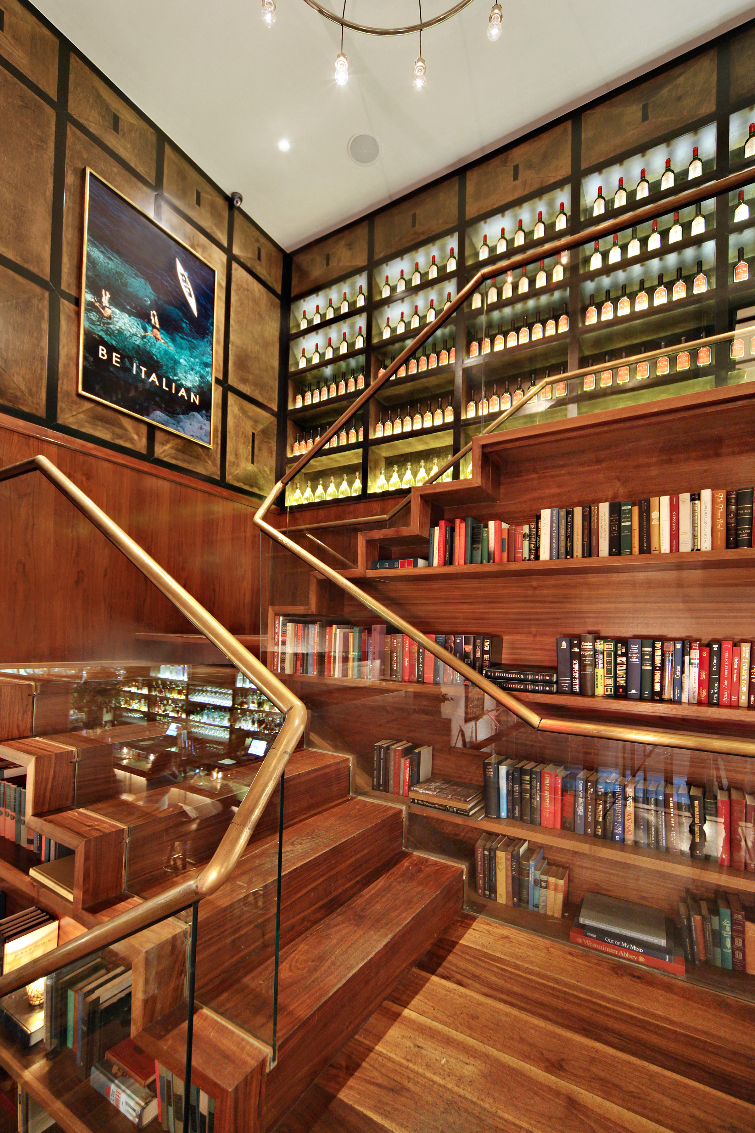 A signature staircase at Felice 15 Gold featuring warm wood finishes, glass railings, built-in bookshelves, and a softly illuminated bottle wall, creating an intimate, library-inspired setting that reflects the restaurant&rsquo;s refined Italian atmosphere.