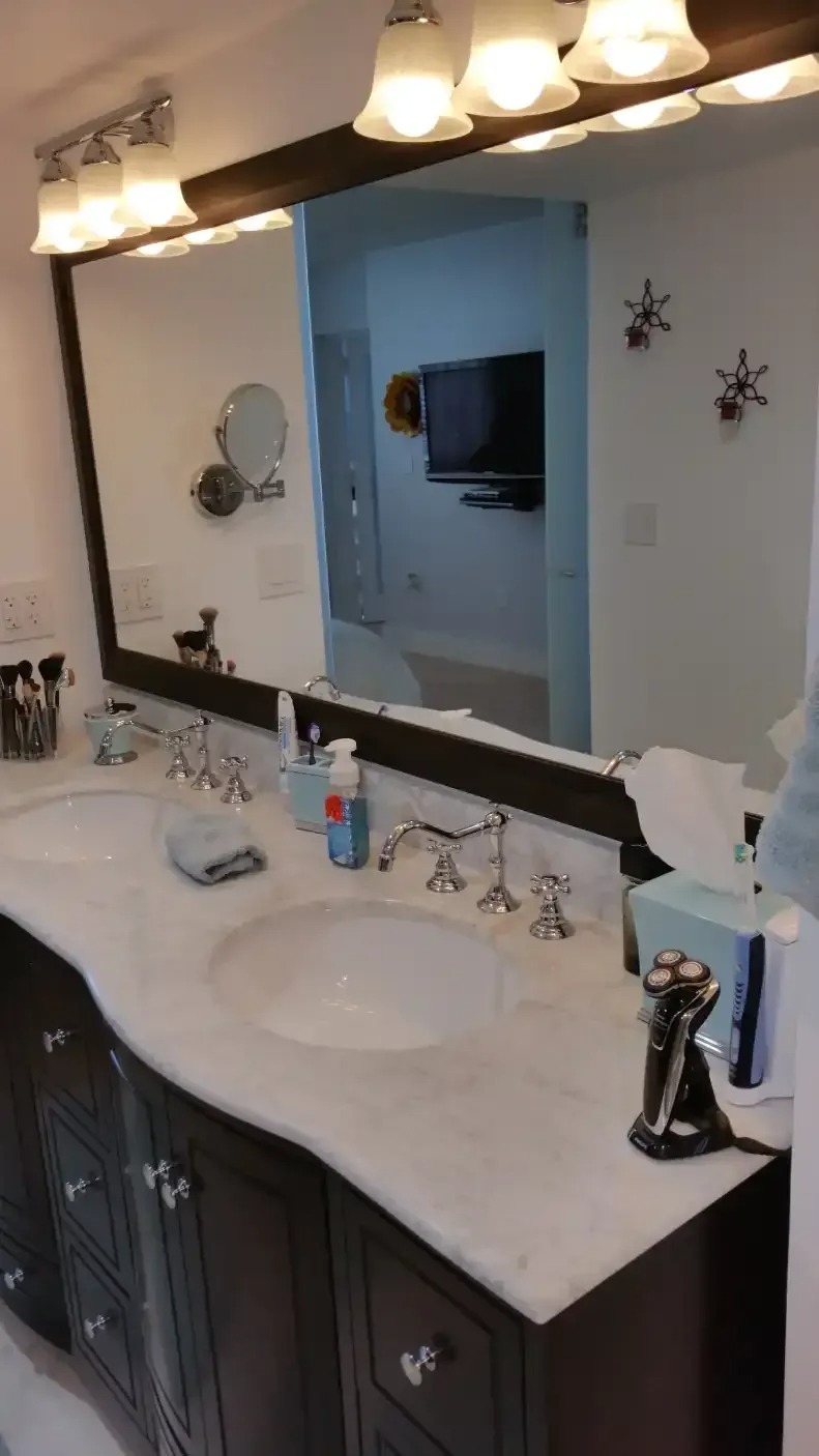 A bathroom features a large, rectangular mirror above a white marble countertop with double sinks. Dark wood cabinets and chrome fixtures are visible. The room is brightly lit.