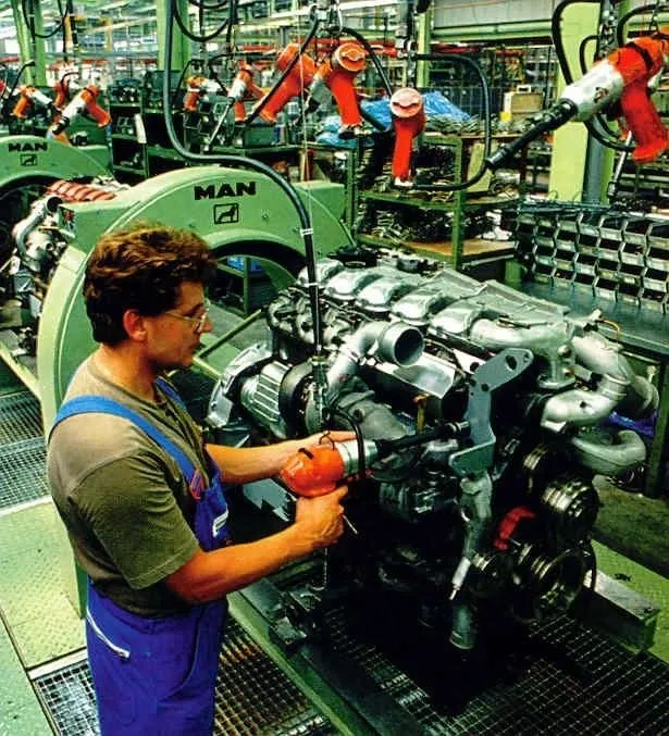 A man in blue overalls and a green shirt works on a large engine assembly line in a factory. He is holding a red power tool and appears to be tightening a component on the engine. Several robotic arms are visible in the background, along with other machinery and engine parts. The factory environment is industrial and busy.