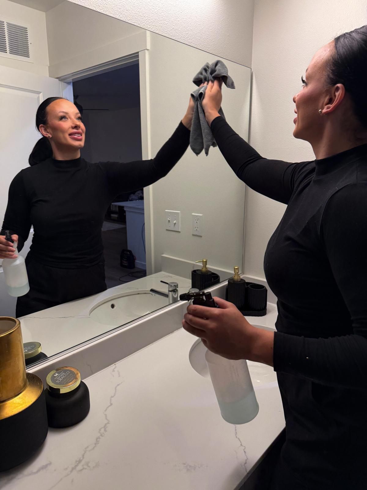 A professional cleaner wipes a bathroom mirror with a cloth while holding a spray bottle, reflecting a clean, modern sink area and demonstrating detailed surface cleaning in a residential space.