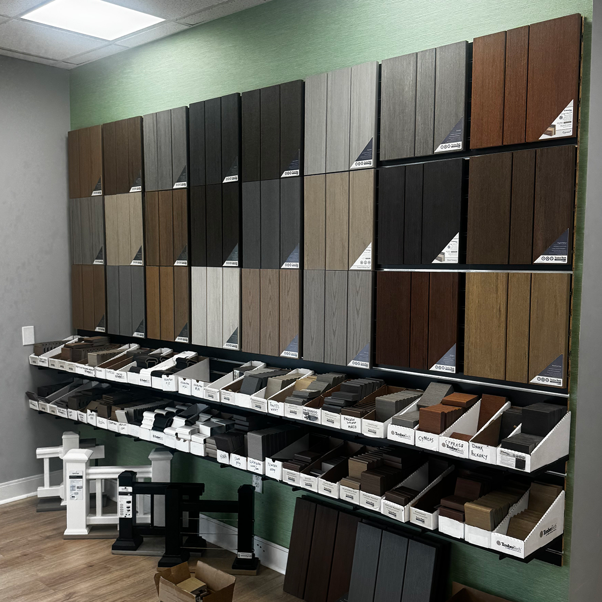 Builders FirstSource showroom wall displaying a selection of decking samples in brown, gray, black and wood-grain finishes. Smaller sample pieces are arranged in labeled boxes on two shelves below, while white and black railing and post samples stand on the floor.