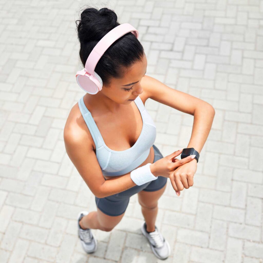 A woman wearing pink headphones and a light blue sports bra is checking her smartwatch while standing on a paved surface.