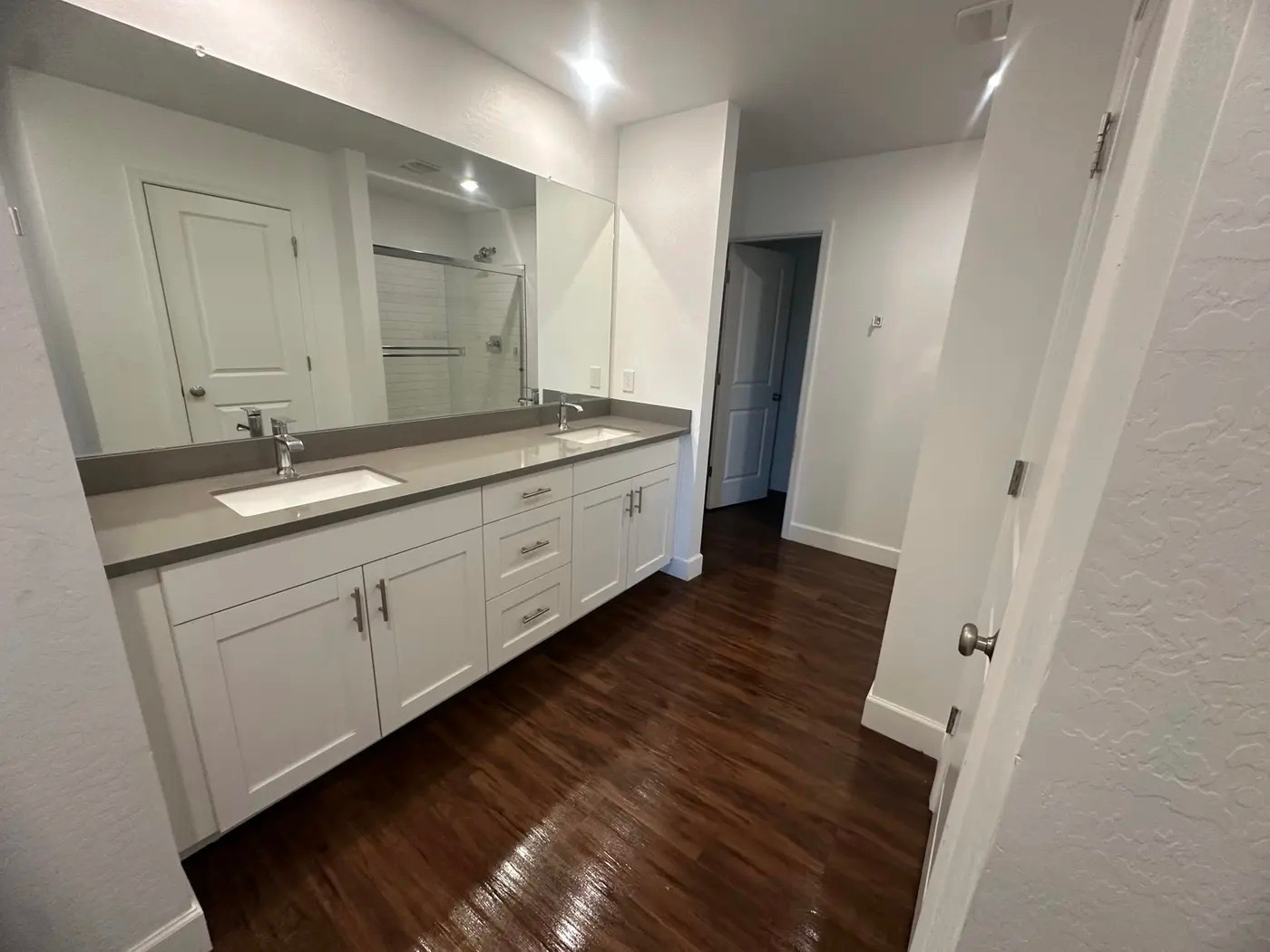 A modern bathroom with a double vanity, large mirror, and a shower visible in the reflection. The room features white cabinetry, a grey countertop, and dark hardwood floors. An open doorway leads to another room.