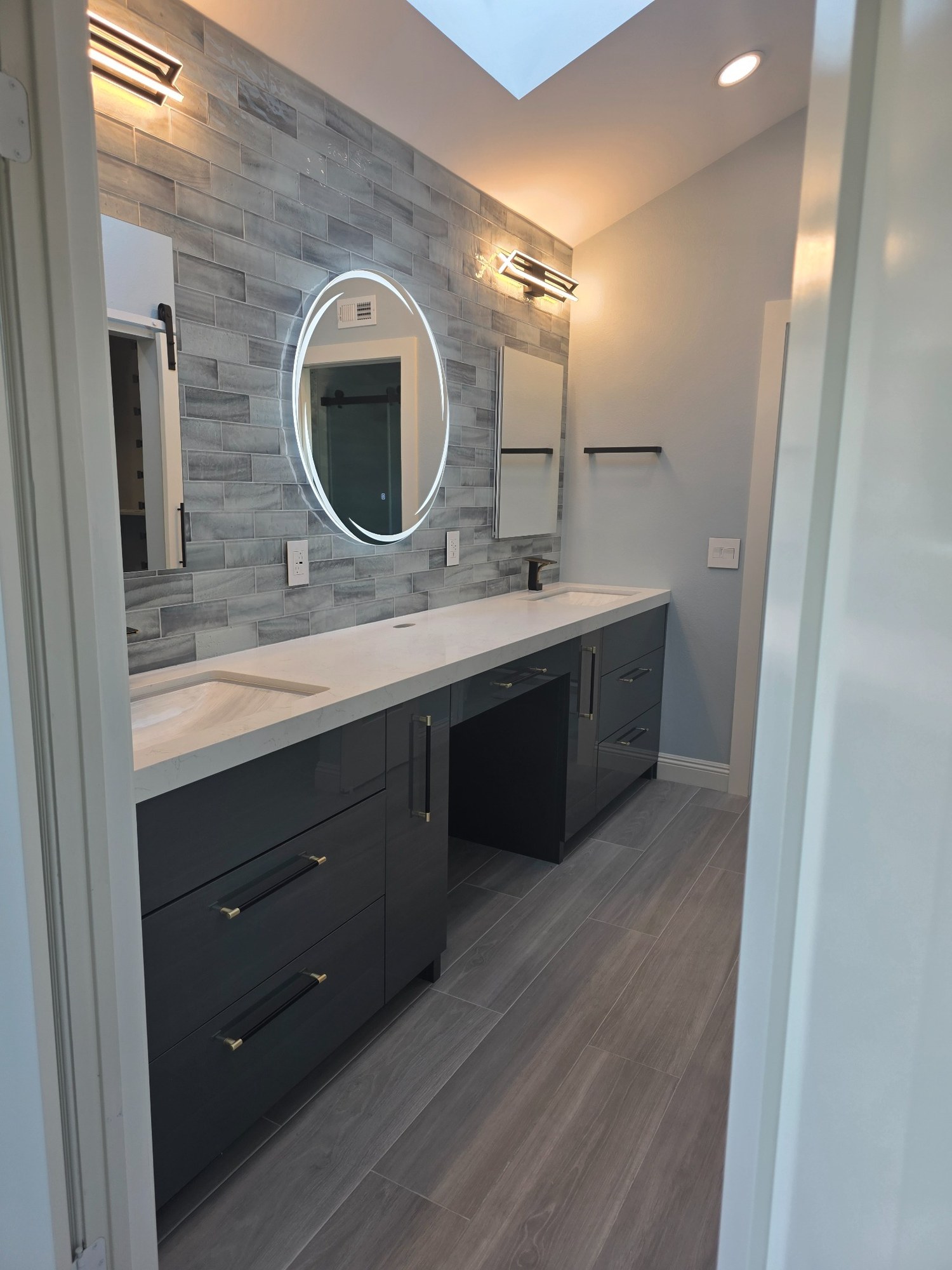 Bathroom vanity remodel with dark cabinetry, stone countertop, round mirror, and wall lighting. 