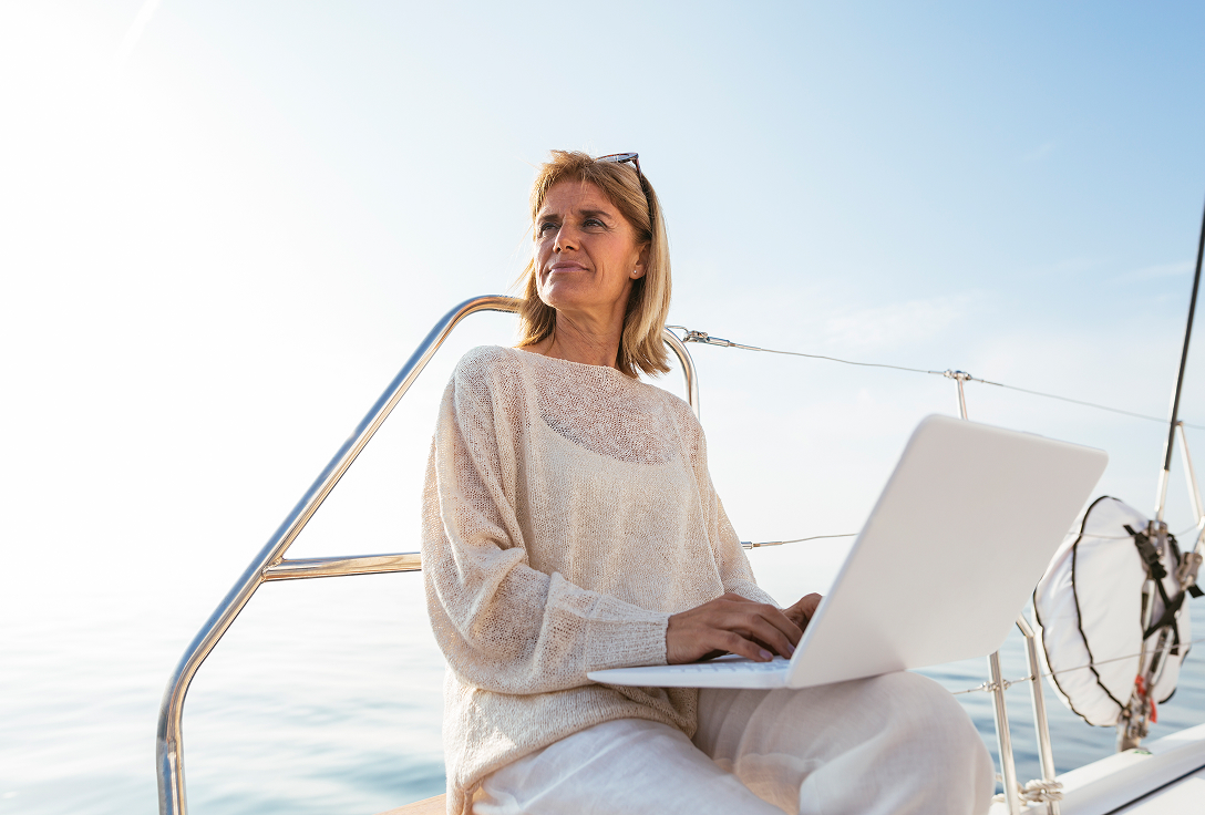 Woman in casual attire works on a laptop aboard a sailboat. She gazes into the distance, conveying relaxation and focus under a clear blue sky.