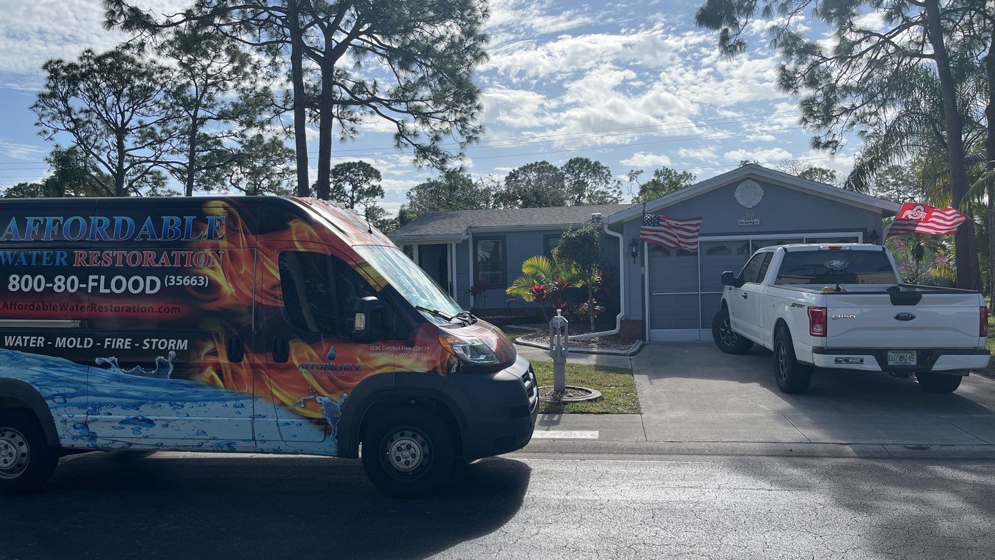 A colorful van from Affordable Water Restoration is parked in front of a house. The van is decorated with flames and water graphics and has text indicating the company's services. A white Ford F-150 pickup truck is parked in the driveway of the house. American flags are displayed on the house.