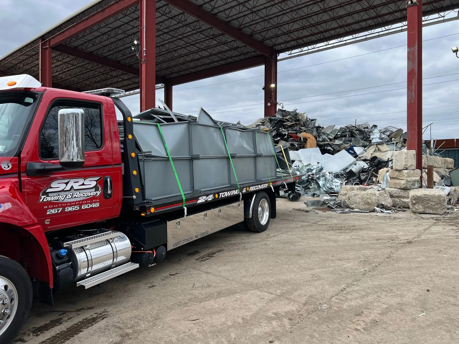 A red SRS Towing & Recovery flatbed truck loaded with large metal scrap, secured with green straps, parked at a scrapyard under a metal canopy with heaps of scrap metal in the background.