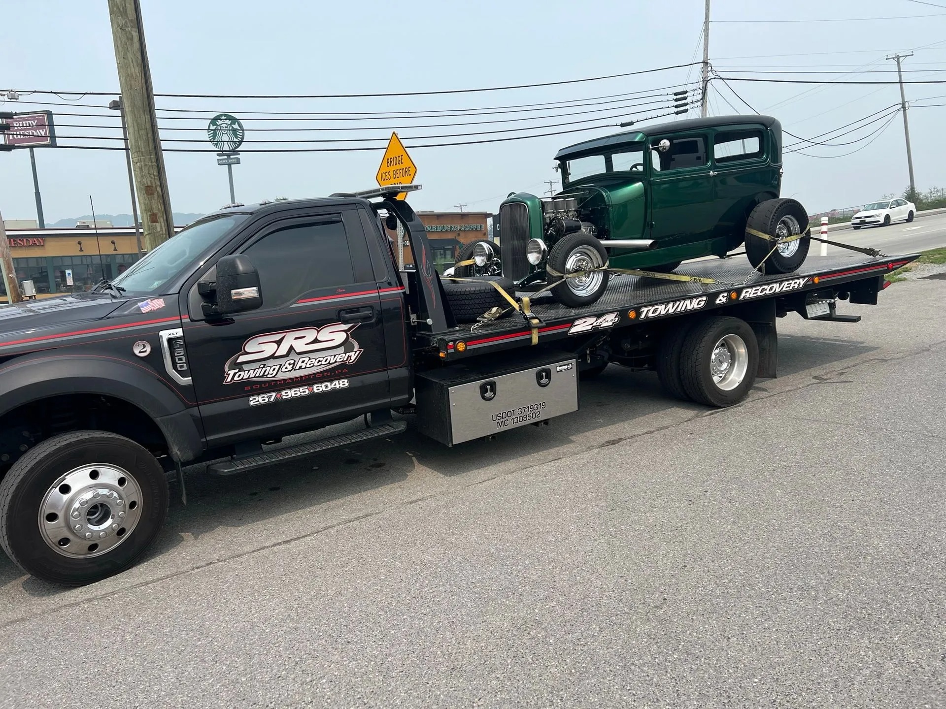 A black SRS Towing & Recovery tow truck transporting a vintage green hot rod on its flatbed, secured with yellow straps.