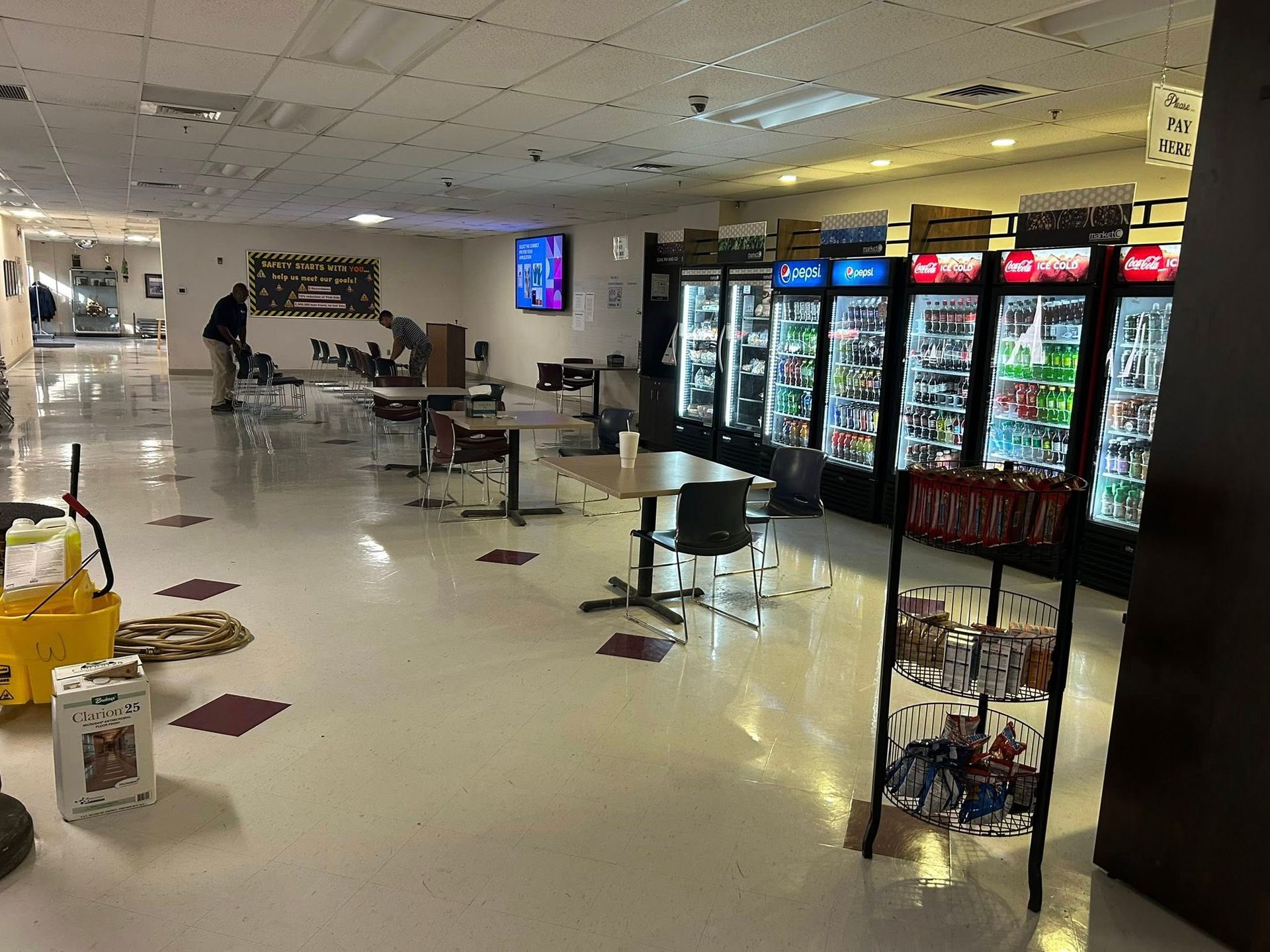 Commercial breakroom or cafeteria with tables, chairs, beverage coolers, and vending displays, while workers clean and arrange seating, showcasing professional janitorial services maintaining cleanliness and organization in a workplace environment.