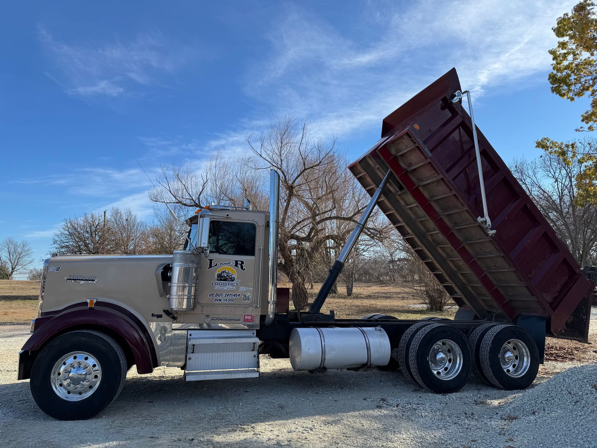 L & R Logistics LLC silver and burgundy dump truck photographed from the side with its bed raised, showcasing heavy-duty hauling equipment and hydraulic lifting components outdoors locally in Oklahoma.