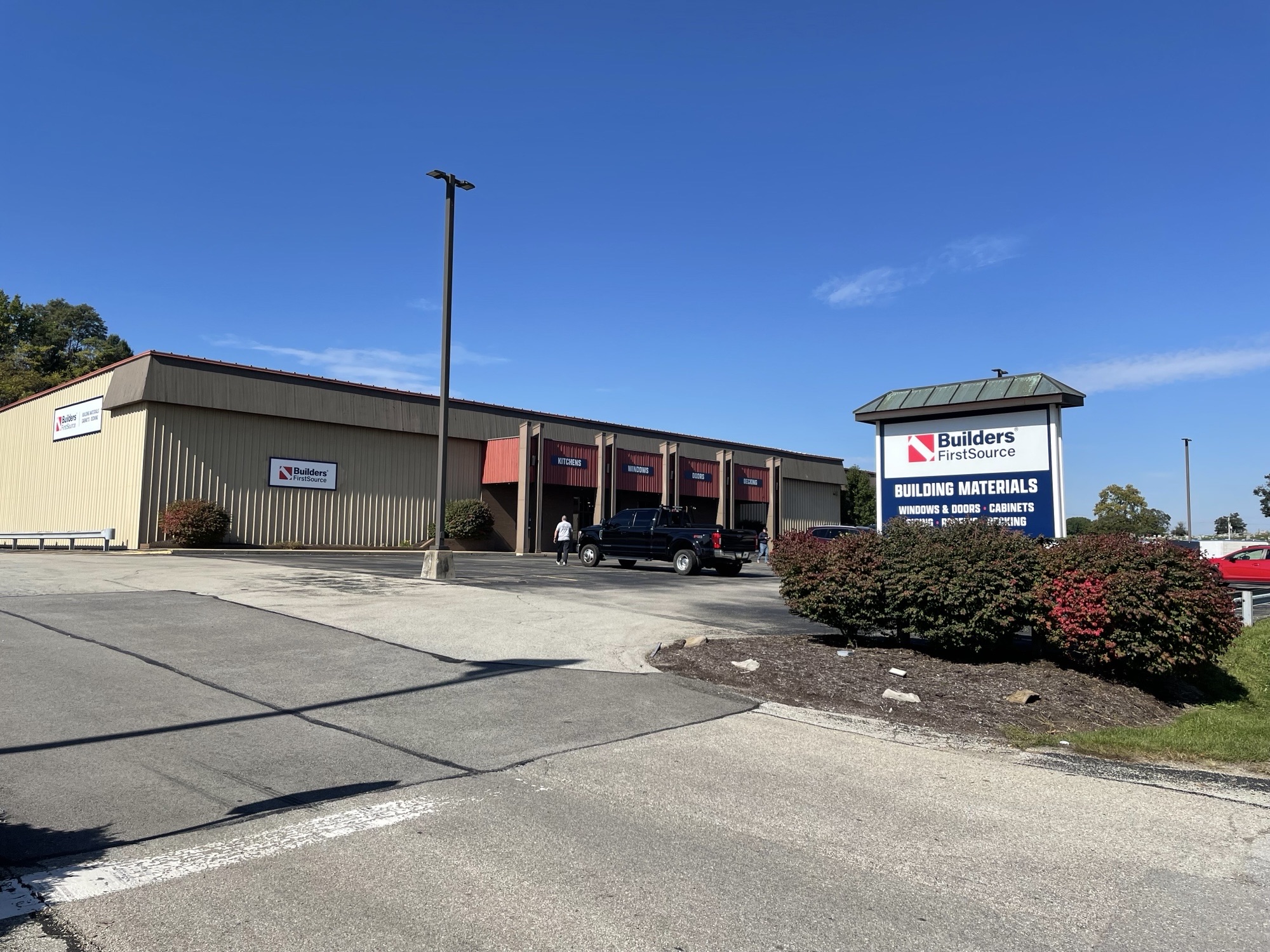 Exterior view of a Builders FirstSource location with a large roadside sign advertising building materials, windows, doors, cabinets, and decking. The building sits beside a parking lot under a clear blue sky.
