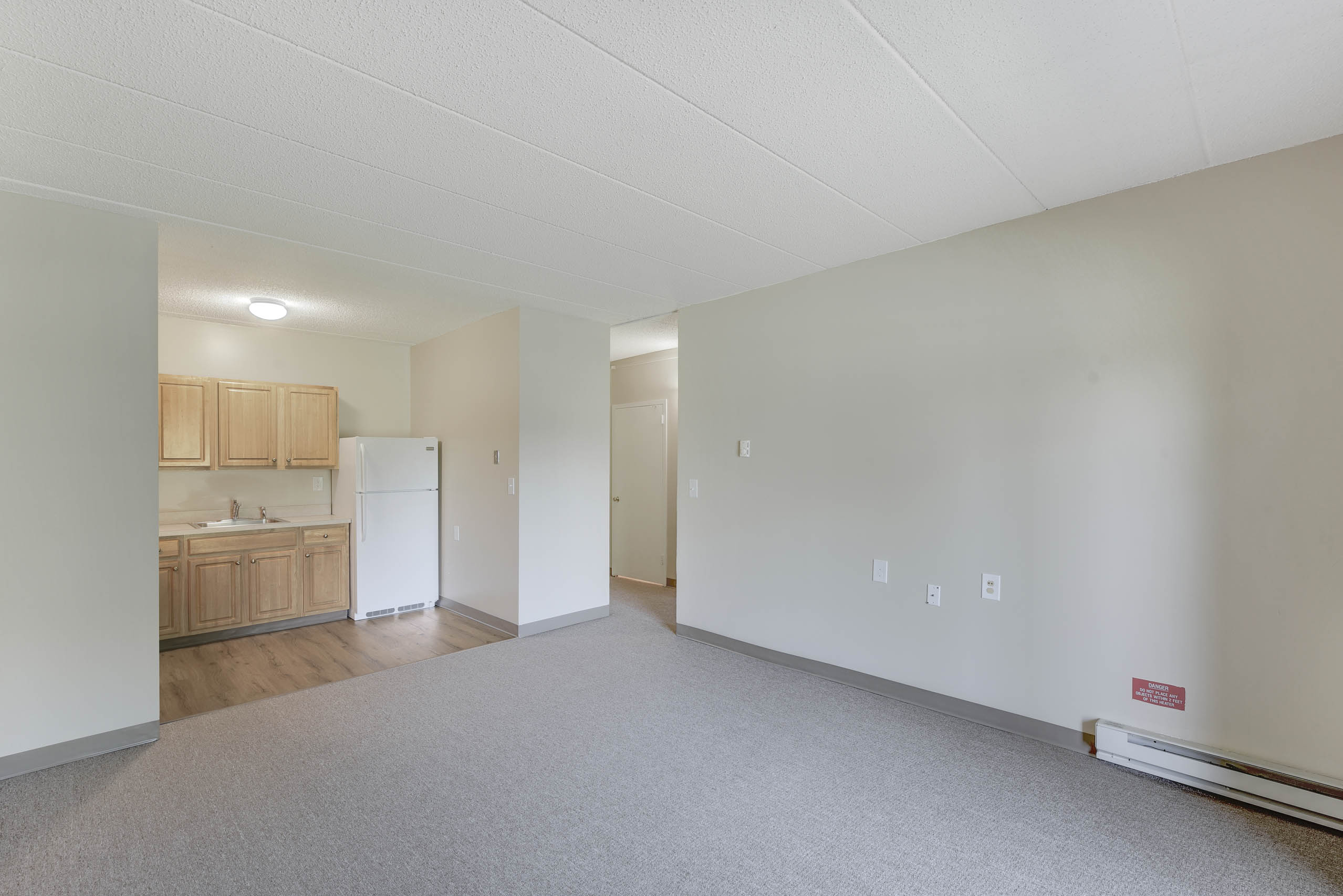 A large empty room with a kitchen area visible through the door.