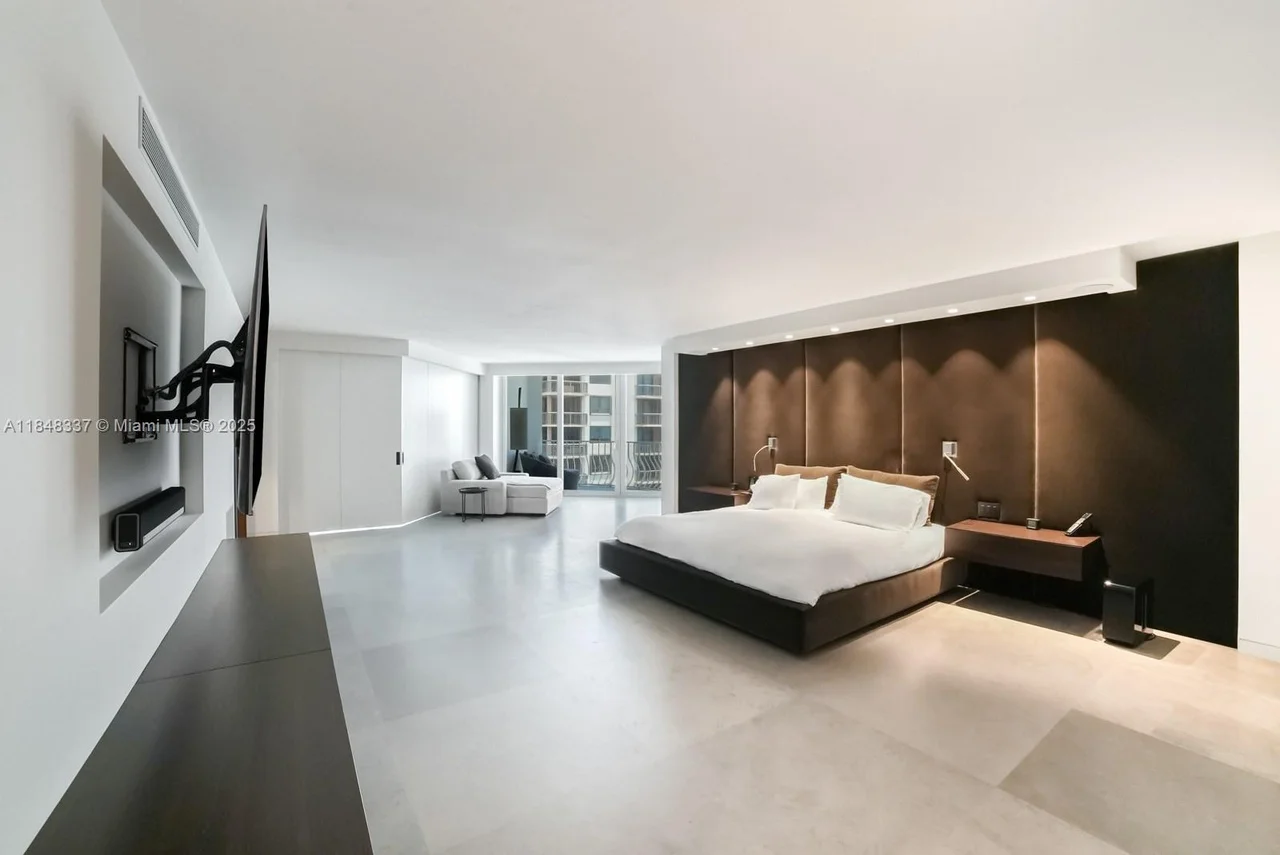 A spacious bedroom with a large bed and a seating area in the background. The room features modern decor with a large dark brown upholstered headboard, a wall-mounted television, and a minimalist design.