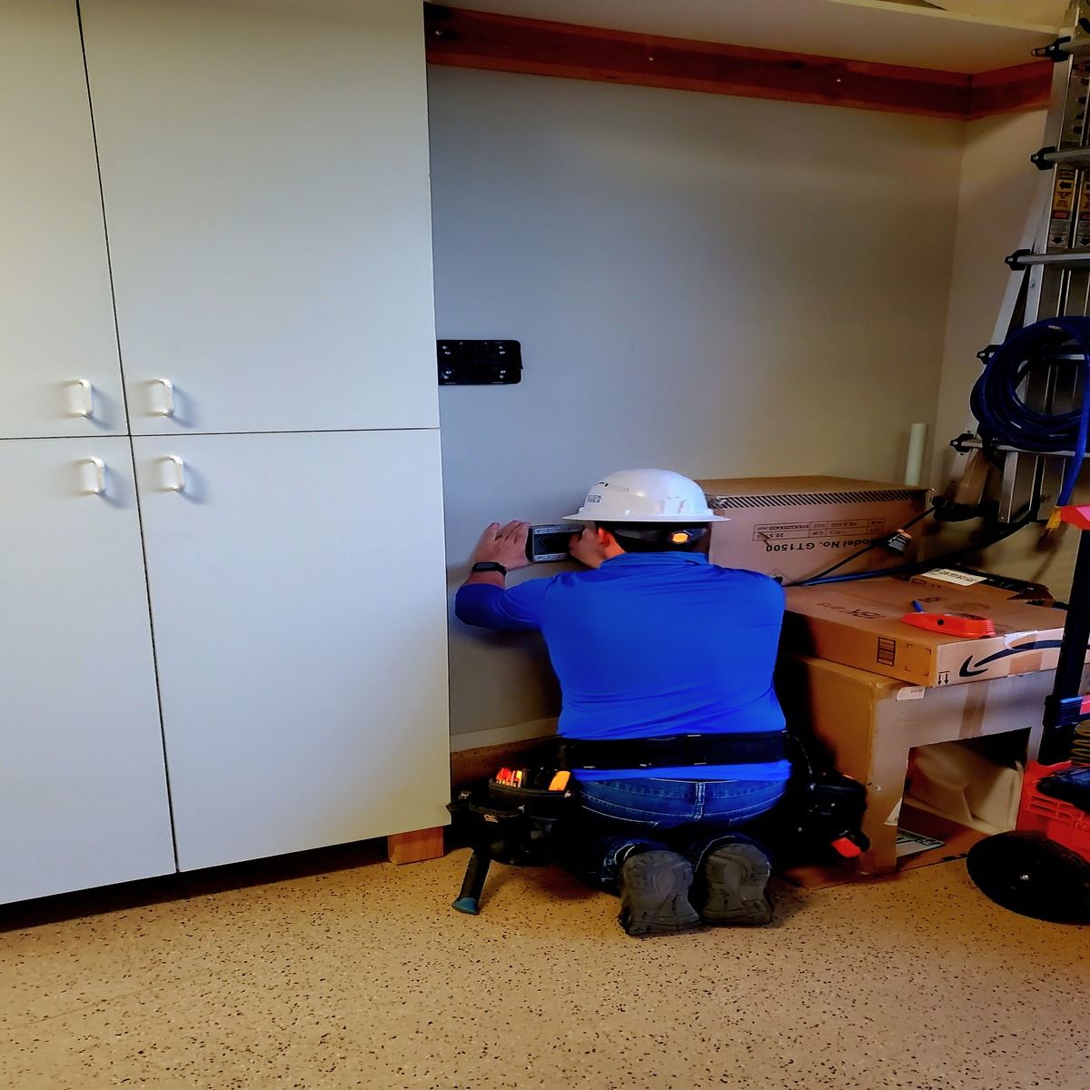 An electrician kneels inside a residential garage while mounting electrical equipment on the wall beside storage cabinets, boxes, tools, and a ladder during an electric vehicle charger installation project indoors.
