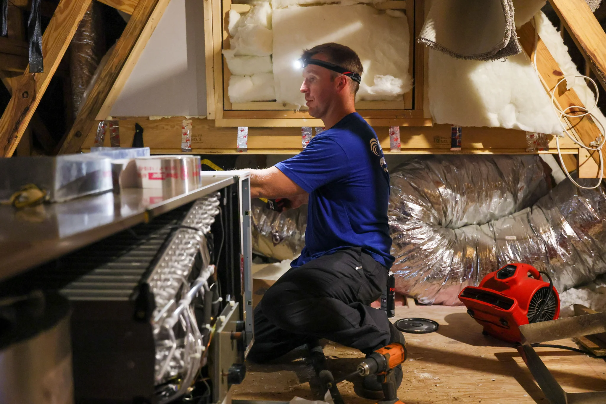 A man wearing a blue t-shirt and black pants with a headlamp is working in an attic. He is kneeling  