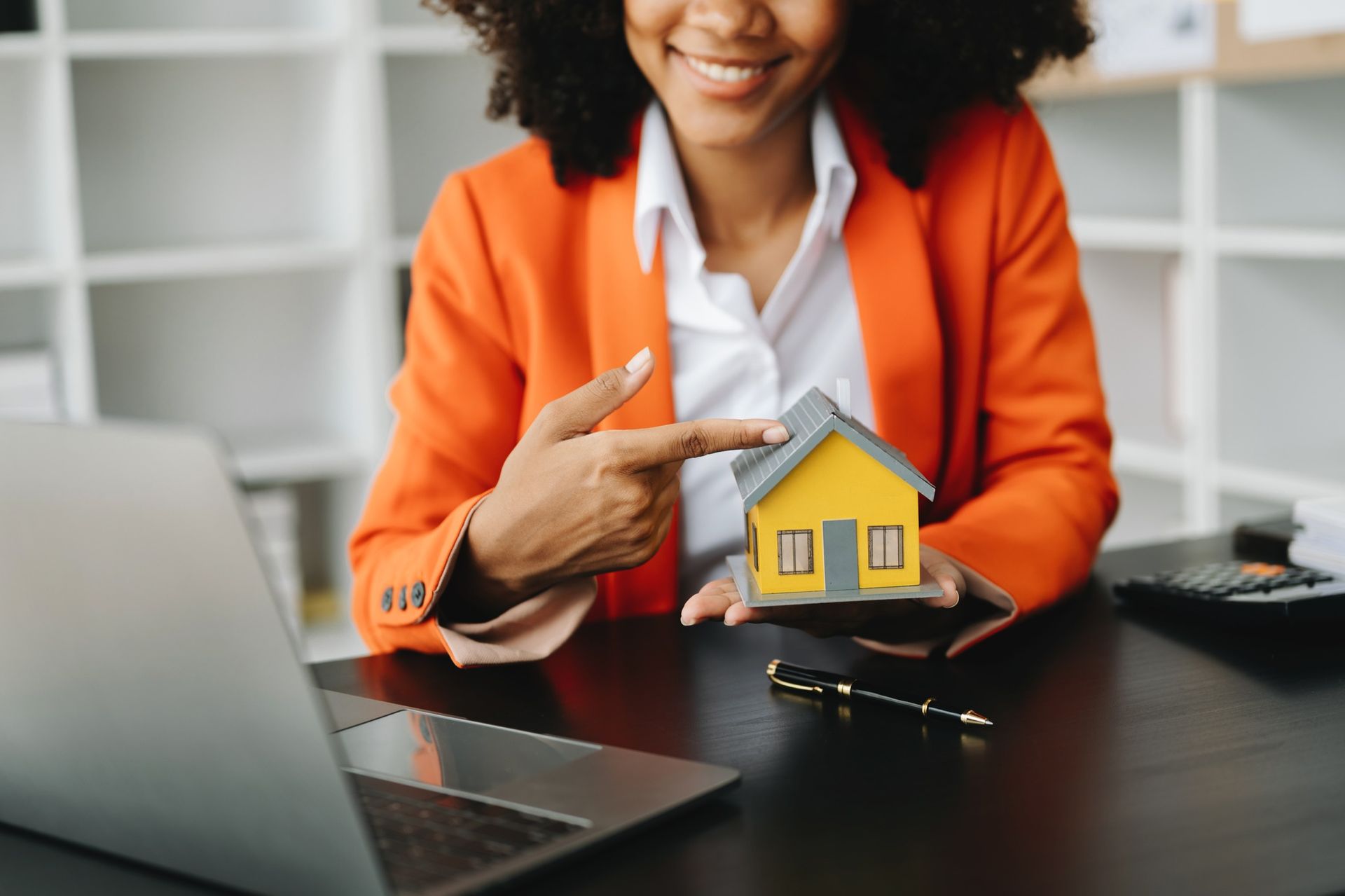 Professional real estate consultant presenting a miniature house model while sitting at an office desk. The image reflects property advisory services, homeownership guidance, real estate investment consulting, and customer-focused support for residential and commercial property buyers seeking trusted expertise.
