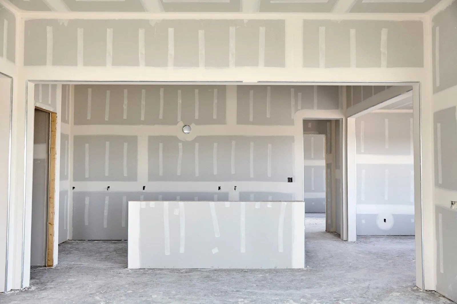 An interior room under construction shows newly installed gray drywall panels. The seams are taped and spackled with white joint compound, ready for final sanding and painting.