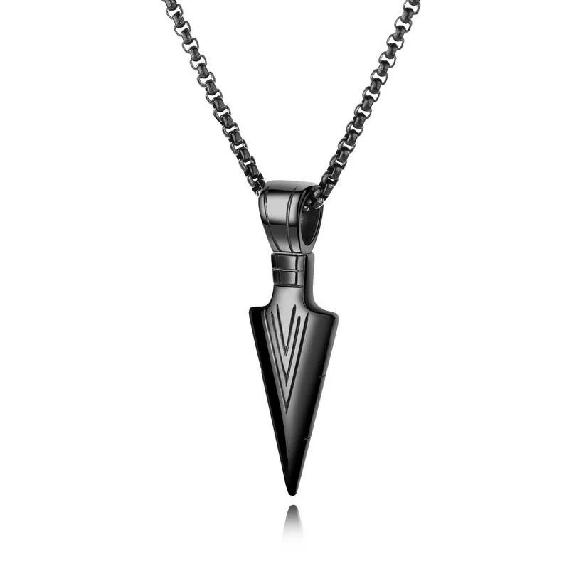 A close-up studio shot of a dark, metallic arrowhead pendant necklace against a white background. The pendant has a matte finish with incised lines forming a V-shape near the tip. The chain is made of dark, interlocking oval links.