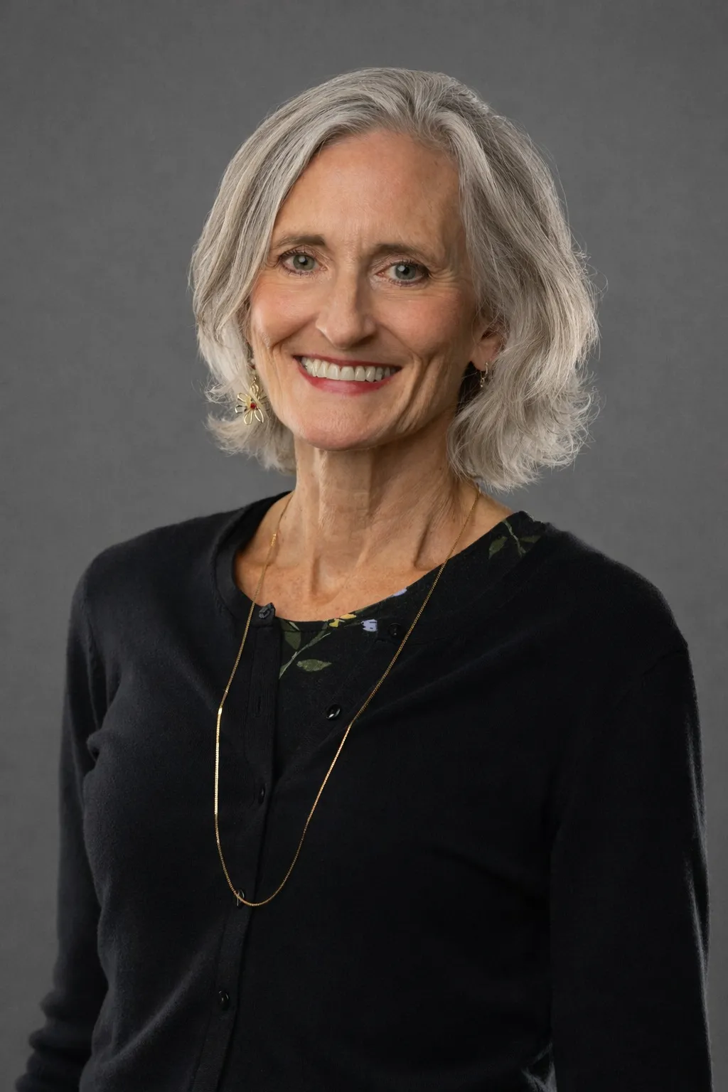 A head-and-shoulders studio portrait of a smiling, middle-aged woman with short, gray hair, fair skin, and blue eyes. She is wearing a black cardigan over a floral print top and a delicate gold chain necklace. Her left earlobe has a dangling earring shaped like a flower with red and gold accents. The background is a plain, dark gray.