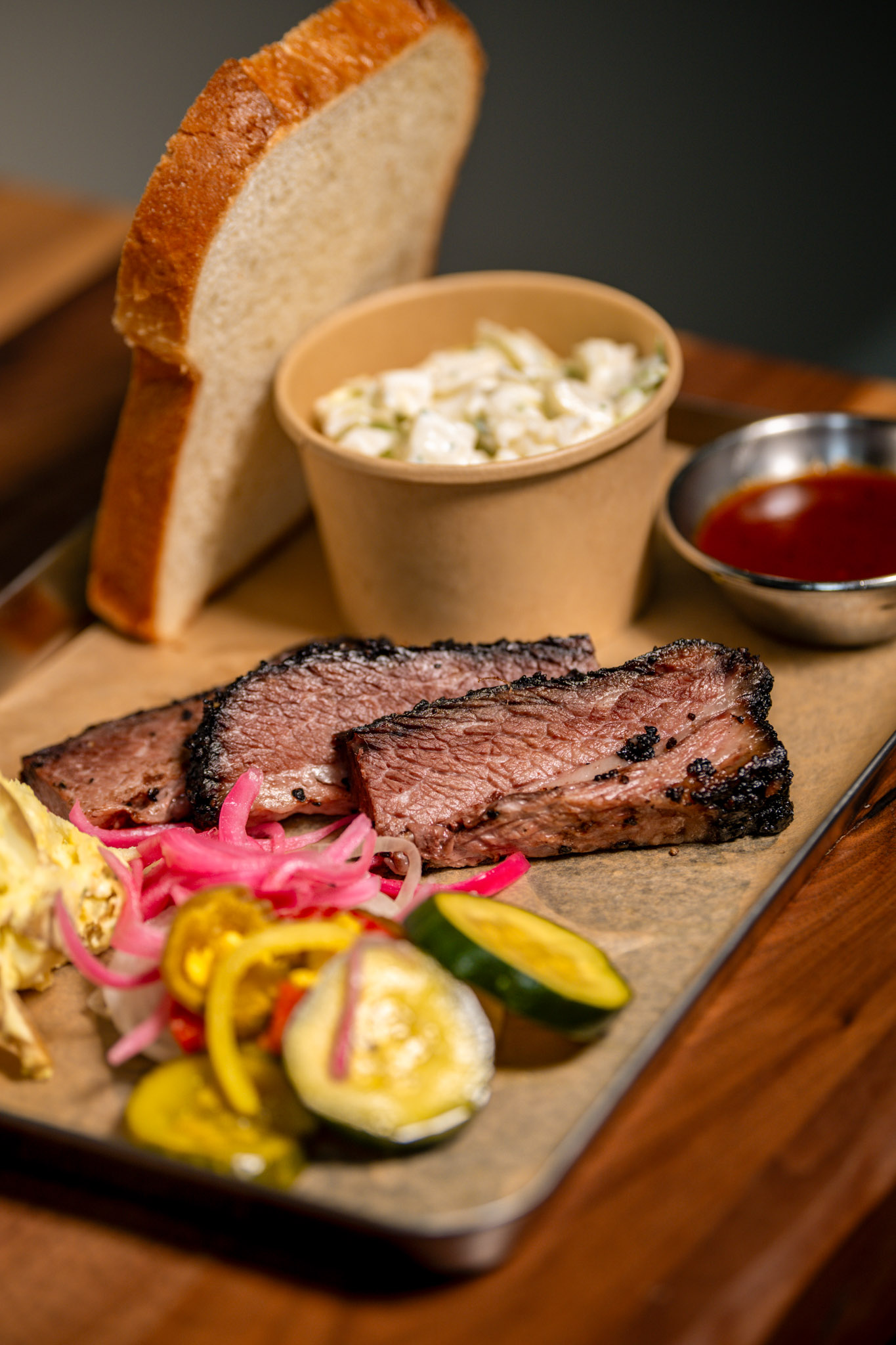 Classic Central Texas Brisket with a deep black pepper crust, served on a metal tray with bread and butter pickles, pickled red onions, white bread, coleslaw, and a ramekin of Joe's BBQ sauce.