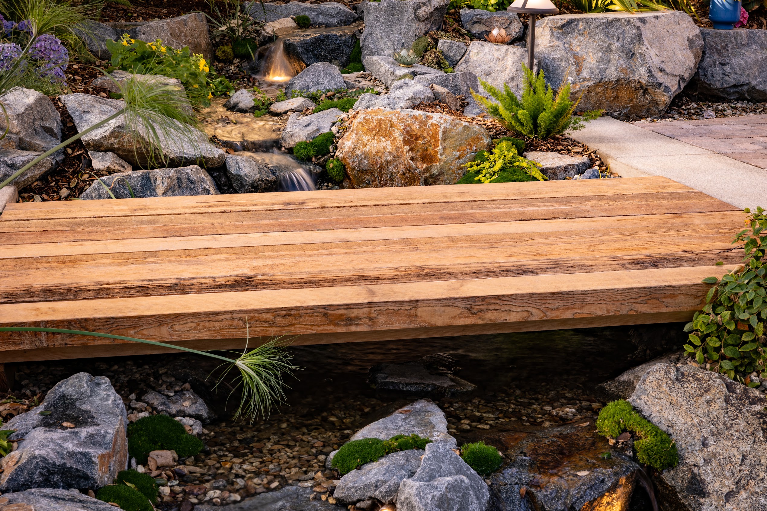 Rustic wooden footbridge spans a clear, rocky stream in a landscaped garden, with waterfalls, boulders, moss, and colorful plants.