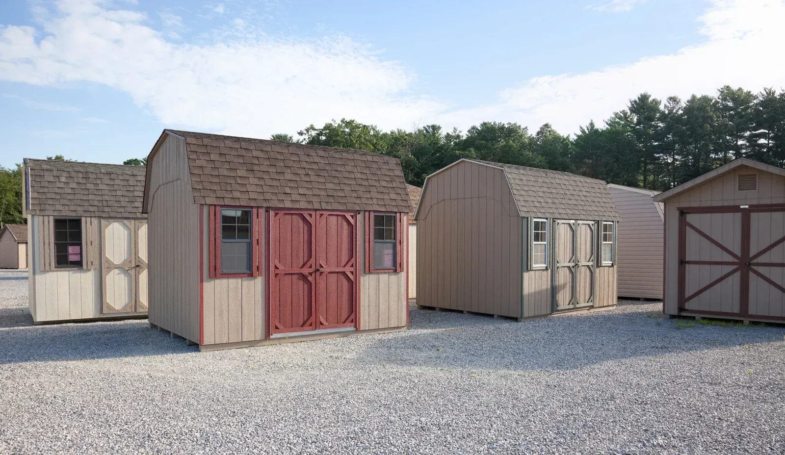 An outdoor display lot showing several pre-built, gambrel-roofed storage sheds on a gravel surface. The shed in the foreground has light tan siding with dark red trim and barn-style double doors, flanked by windows.
