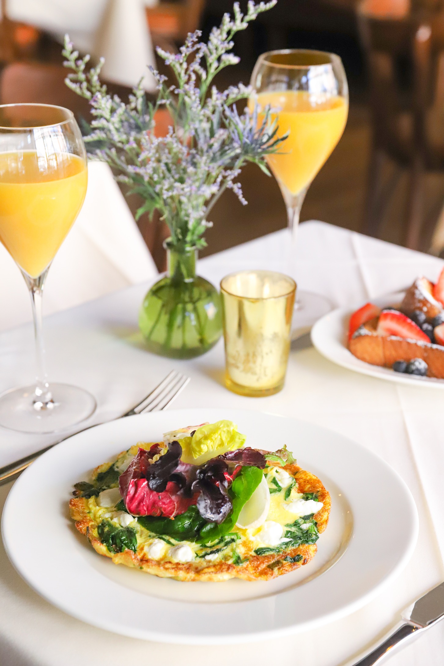 A vibrant morning meal featuring a vegetable-heavy omelet topped with mixed greens, served with chilled mimosas and fresh floral accents.