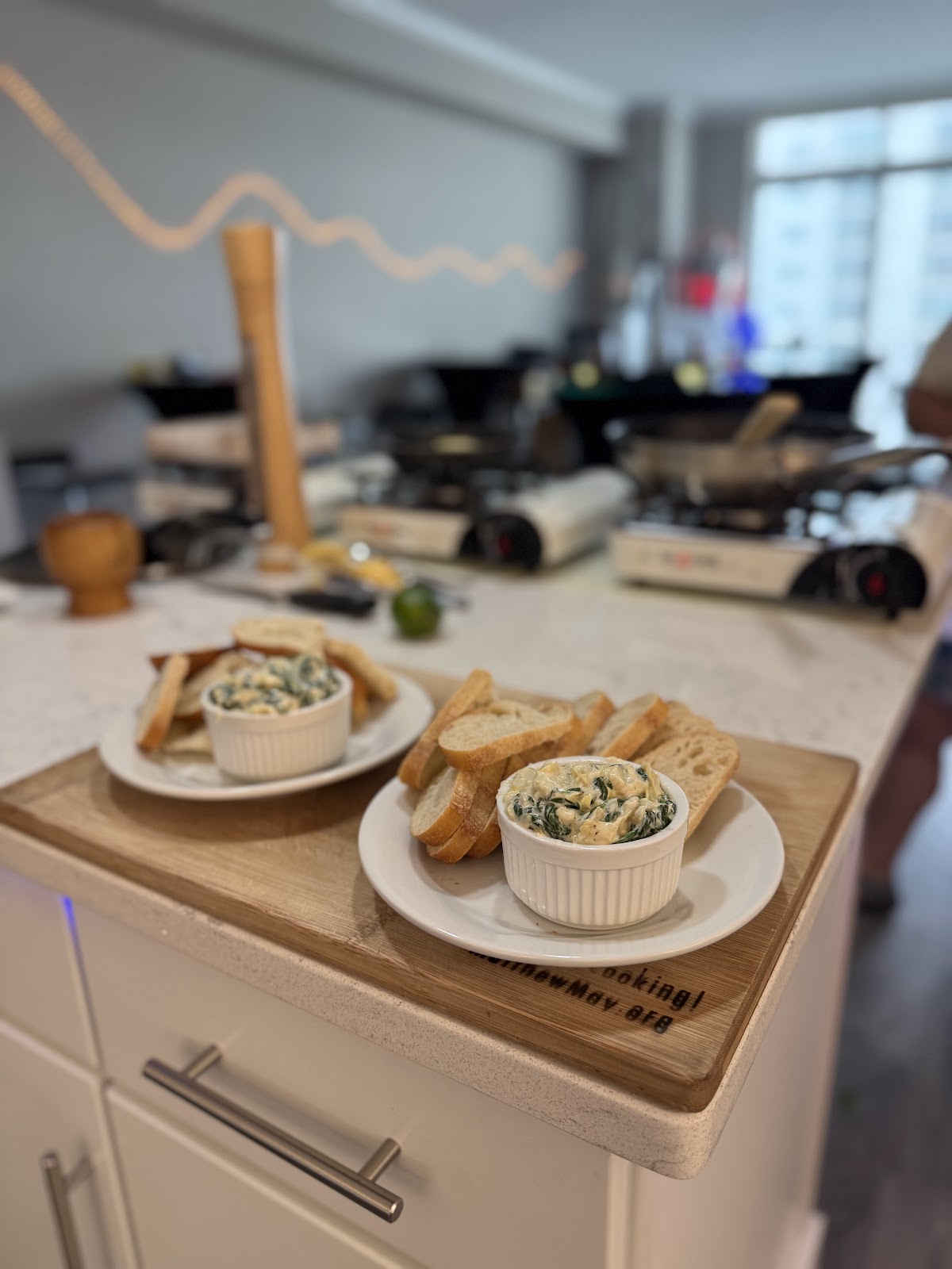 Two white ramekins filled with a creamy green dip, possibly spinach or artichoke dip, are served with slices of toasted baguette on white plates. The plates are arranged on a wooden cutting board, which rests on a white countertop. In the background, a kitchen counter is visible with multiple portable butane stoves, frying pans, and other kitchenware, suggesting a cooking or dining event in progress. The lighting is bright, with some decorative string lights visible in the upper background.