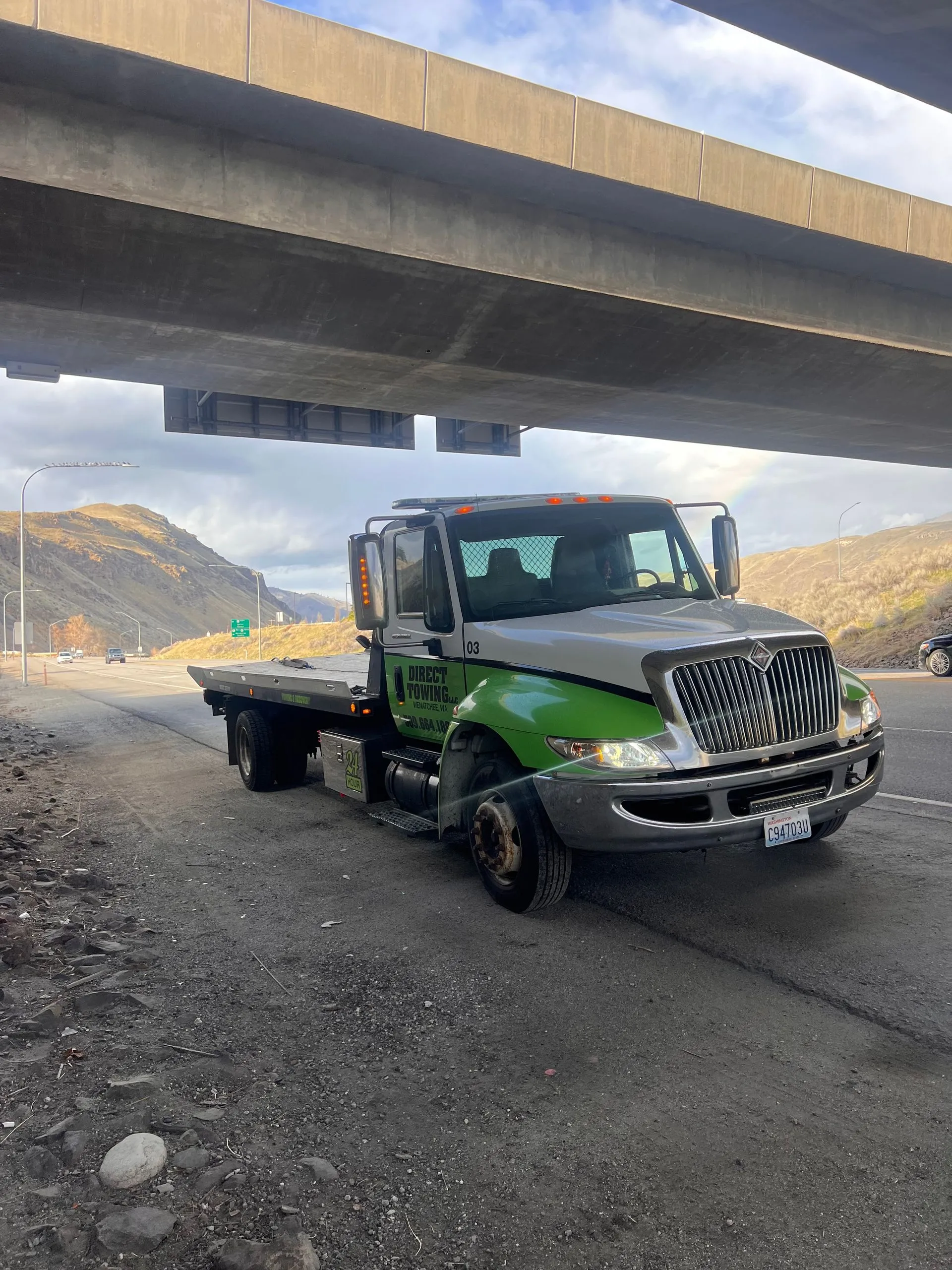 Direct Towing flatbed truck parked beneath a highway overpass with mountain scenery in the background, representing roadside assistance, vehicle recovery services, and dependable transportation across regional routes.