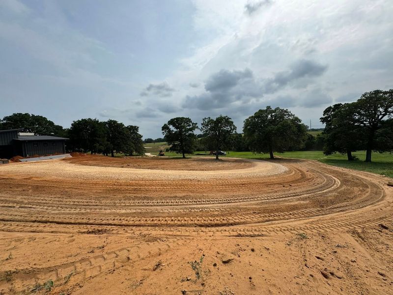 Freshly graded circular dirt driveway or track surrounding property, showcasing precision grading, site preparation, and land shaping services for residential development with smooth finish and defined edges in natural setting.