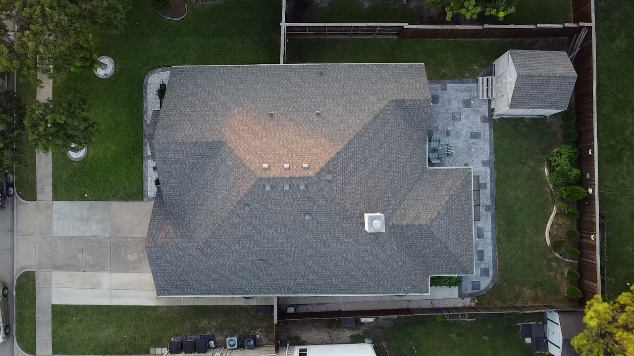 An aerial view of a residential property showing a large house with a gray shingled roof, a paved driveway, a backyard patio with chairs, a small shed, and surrounding green lawns and trees. The scene is captured during daylight.
